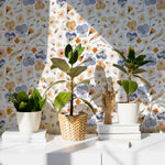 A vibrant and artistic display of the Blooming Boho Floral Wallpaper in a room setting. The wallpaper is adorned with a pattern of blue and orange flowers interspersed with subtle green leaves. The scene includes potted plants and a wooden hand model on a white cabinet, highlighting the wallpaper's compatibility with natural decor elements.