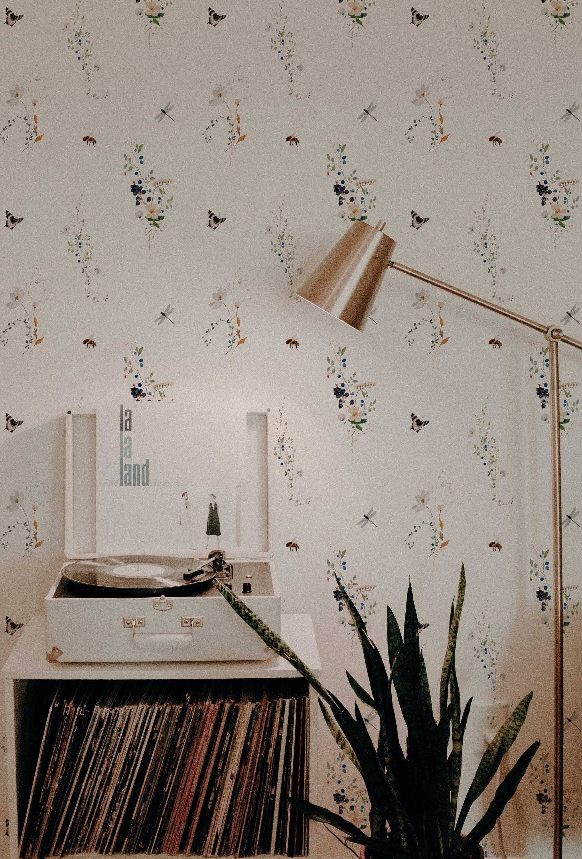 A cozy corner with a playful and botanical themed wallpaper showing a variety of insects and small flowers. This setting includes a record player on a white stand, next to a large green houseplant, enhancing the room's vintage and natural aesthetic.