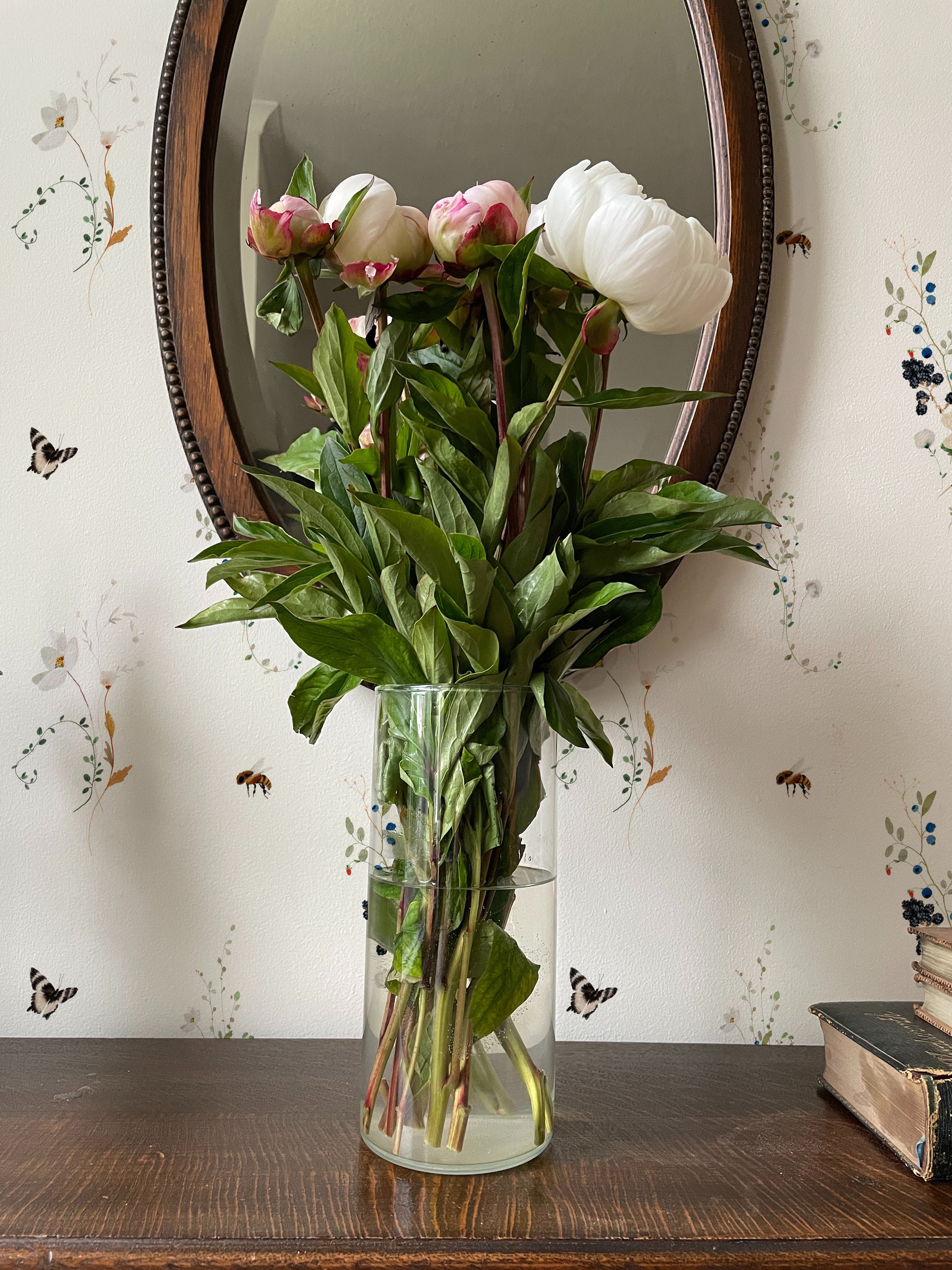 A rustic and charming interior featuring a vintage round mirror with a bouquet of pink and white tulips in a clear vase set on a wooden surface. The wall behind is adorned with a delicate wallpaper depicting small flowers, insects, and butterflies, creating a warm and inviting ambiance.