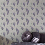 A luxurious bedroom corner featuring the Wisteria Garden Wallpaper, which displays lush clusters of wisteria in shades of purple with green foliage on a light background. The scene is enhanced by the presence of a plush velvet sofa with complementing dark pillows.