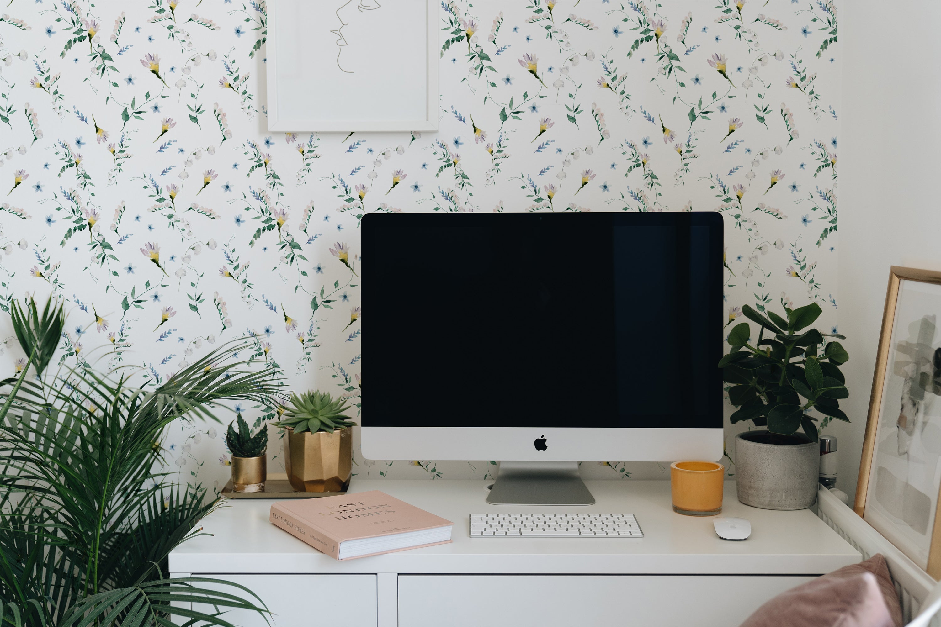 A serene workspace enhanced by the Blossom Breeze Wallpaper featuring delicate floral patterns with tiny blue and yellow flowers and green leaves against a white background. The vibrant natural theme complements the modern desktop setting, surrounded by lush indoor plants and stylish decor.