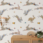 A well-lit nursery featuring the 'Floral Bunnies Wallpaper' as a focal point. The wallpaper's array of leaping and sitting rabbits, decorated with floral prints, complements the minimalist decor, including a white crib and simple furnishings, creating a serene and stylish space.