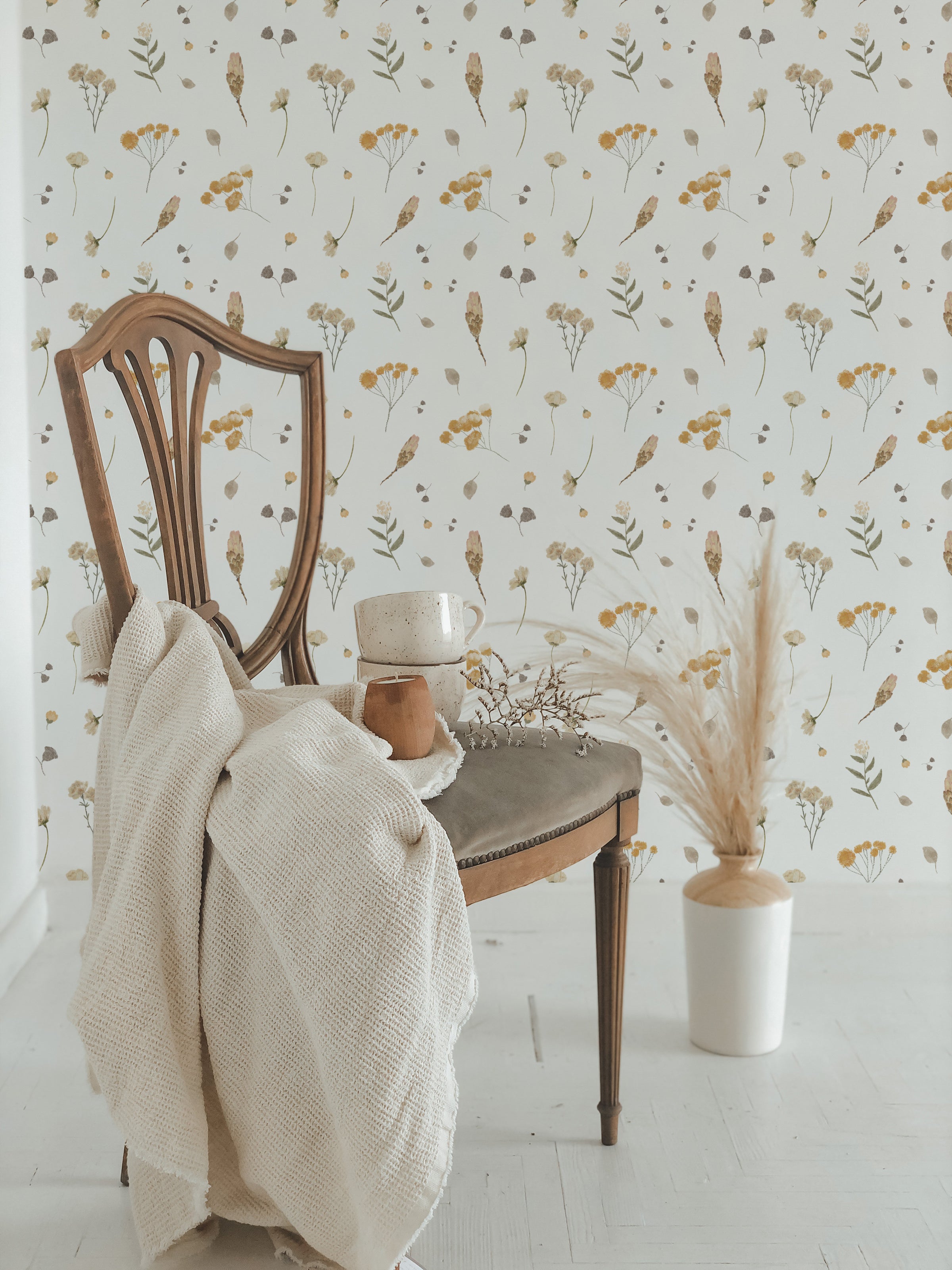 An elegant vintage chair draped with a cozy cream knit blanket sits in front of a wall covered in Buttercup Fields Wallpaper, showcasing a whimsical pattern of yellow buttercup flowers and soft gray foliage on a white background.