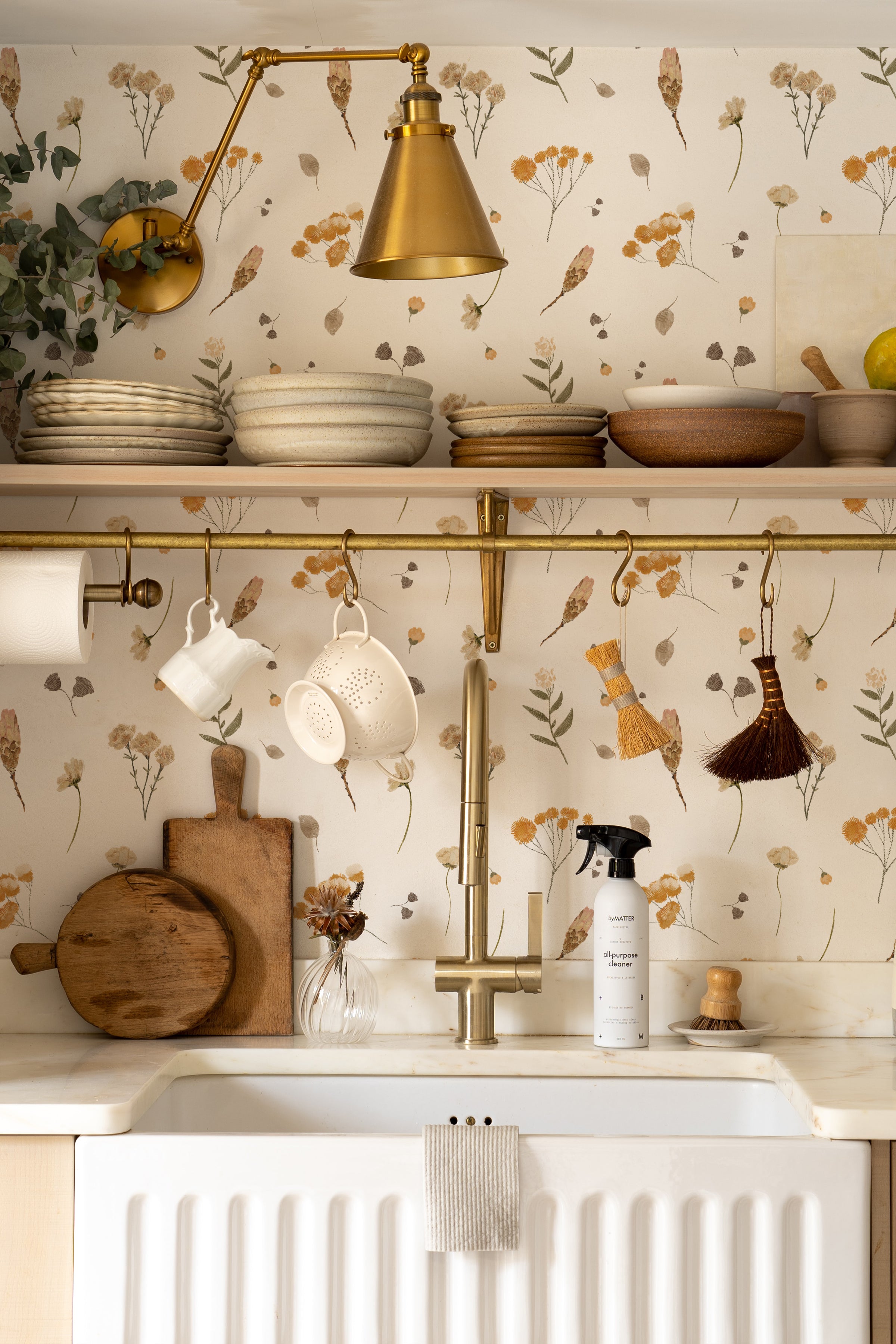 A charming kitchen corner with open shelving displaying ceramics and wooden utensils. The Buttercup Fields Wallpaper lines the wall behind a farmhouse sink, providing a cheerful and rustic backdrop with its yellow and gray botanical print.
