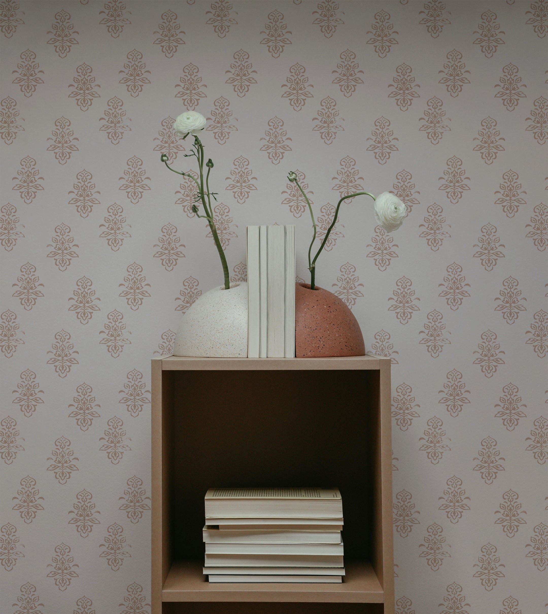 An interior scene featuring the Antique Chic Wallpaper. The wallpaper's pattern of terracotta motifs on a light beige background provides a stylish backdrop. A small wooden shelf holds decor items, including two vases with long-stemmed flowers and a stack of books, creating a sophisticated and serene setting.