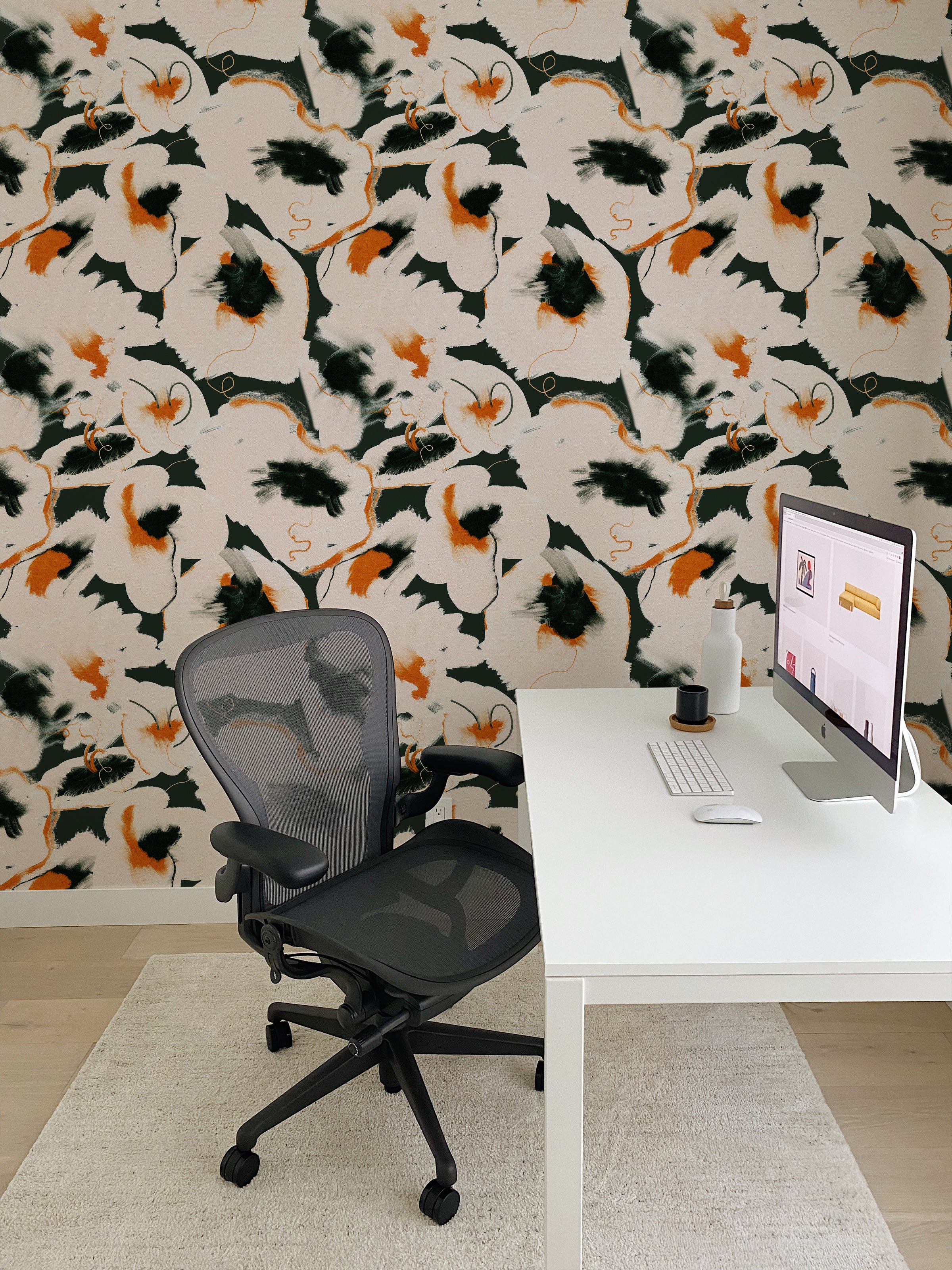 An office space decorated with Abstract Fields Wallpaper, which exhibits a lively abstract pattern with splashes of black and orange on a beige backdrop. The office setup includes a modern desk with a computer, a comfortable office chair, and a cozy white rug, creating a stimulating and creative workspace.
