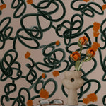 An artistic workspace enhanced by the Abstract Scribble Wallpaper, displaying a unique pattern of tangled green scribbles and orange floral accents on a peach backdrop. The room includes a white chair, a decorative vase with vibrant tulips, and books, creating an inspiring and creative environment.
