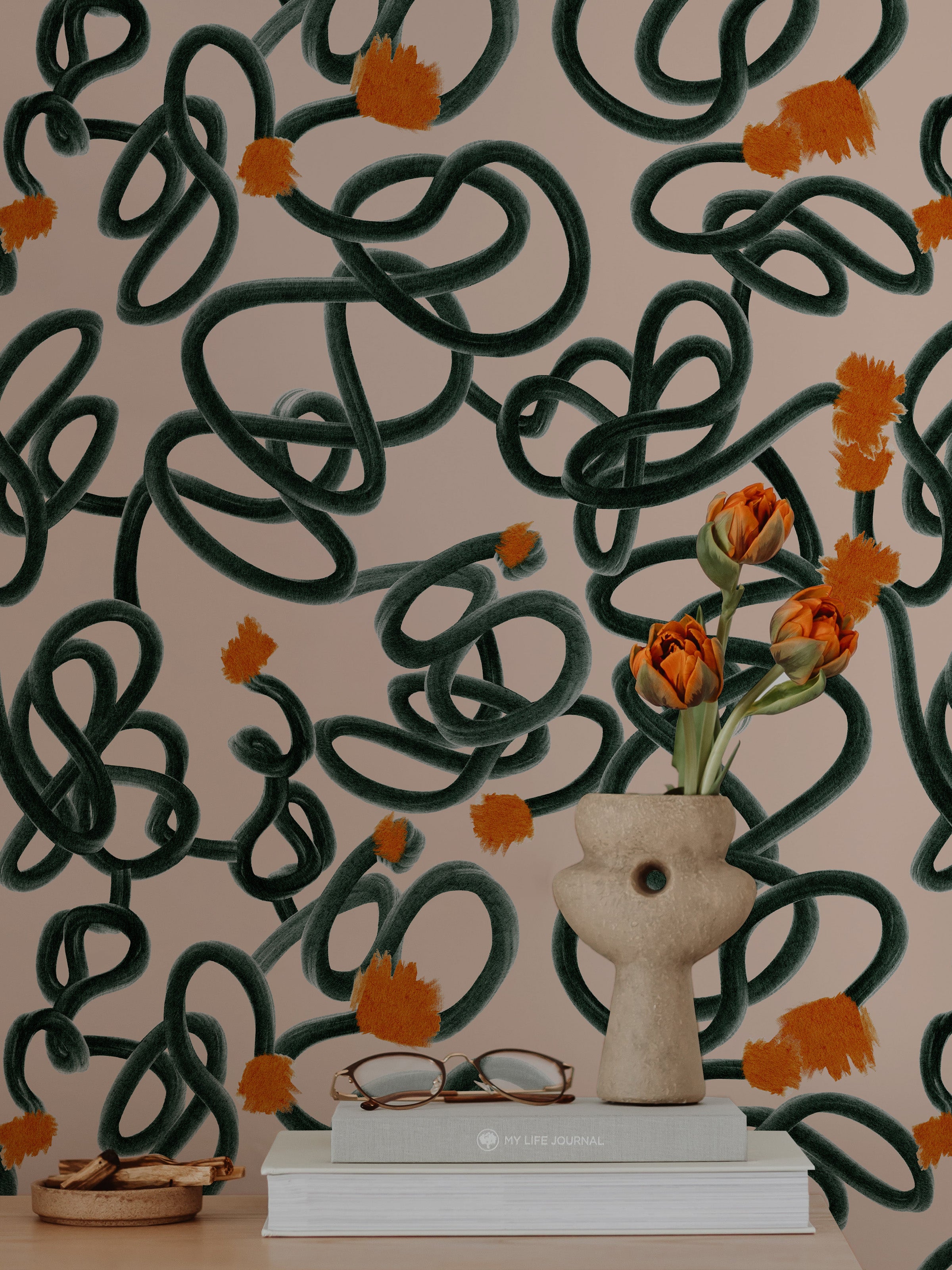 An artistic workspace enhanced by the Abstract Scribble Wallpaper, displaying a unique pattern of tangled green scribbles and orange floral accents on a peach backdrop. The room includes a white chair, a decorative vase with vibrant tulips, and books, creating an inspiring and creative environment.