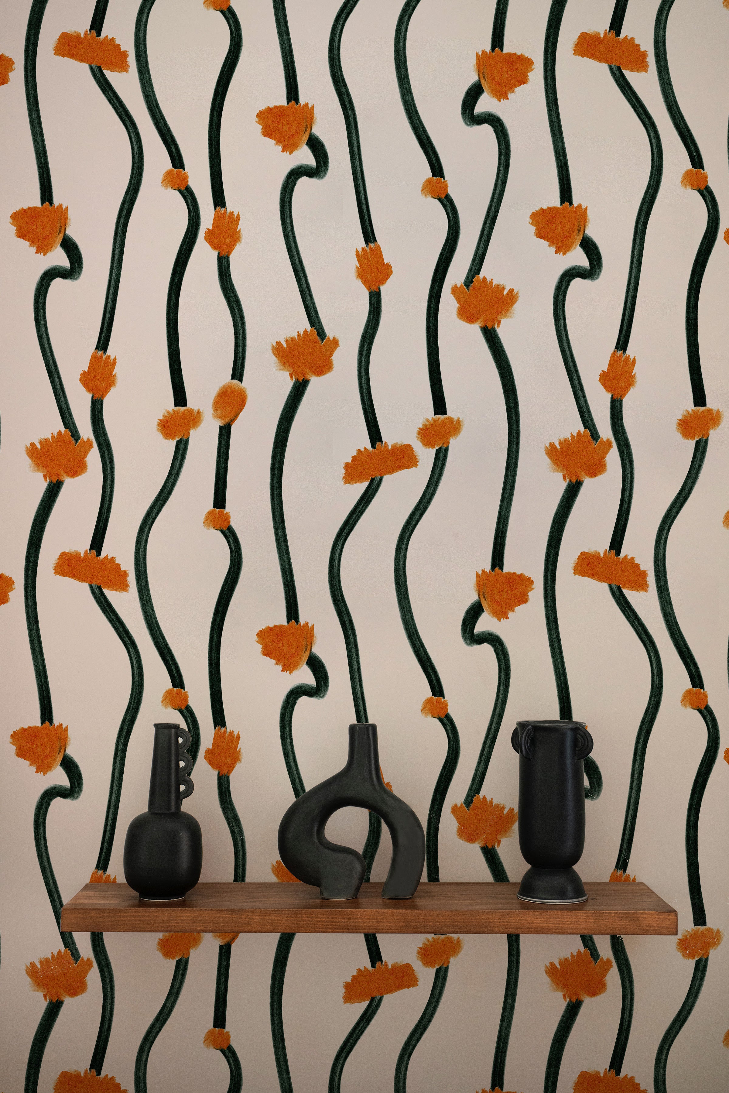 A realistic room setup displaying the same dark green vine and burnt orange floral burst patterned wallpaper. There are two unique black vases placed on a wooden shelf against the wallpaper, enhancing the room's aesthetic.