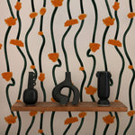 A realistic room setup displaying the same dark green vine and burnt orange floral burst patterned wallpaper. There are two unique black vases placed on a wooden shelf against the wallpaper, enhancing the room's aesthetic.