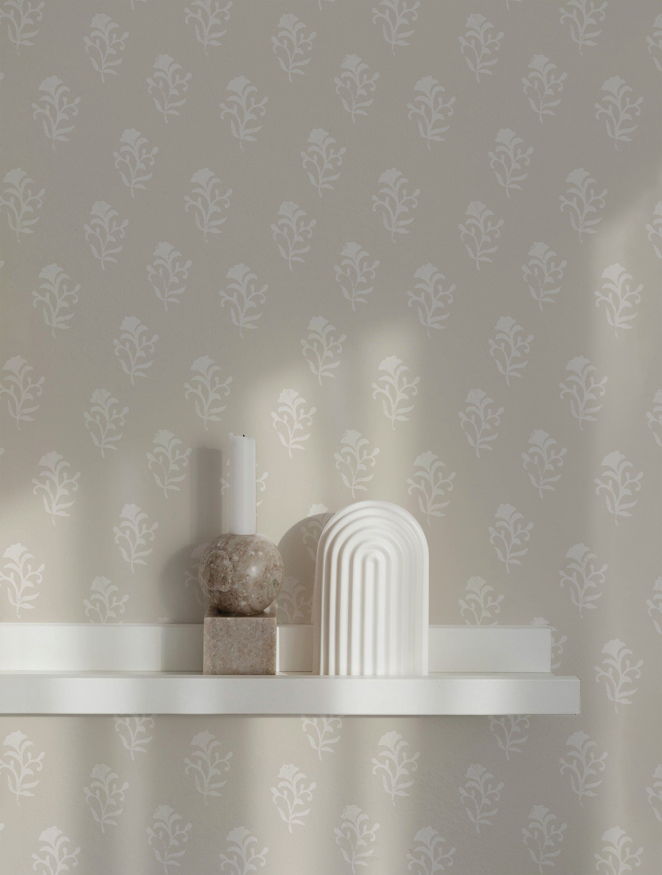 An interior scene featuring the Heritage Fleur Wallpaper. The wallpaper's pattern of white floral motifs on a beige background creates a serene backdrop. A white floating shelf holds minimalist decor items, including a marble sculpture and a white candle holder, illuminated by soft, natural light.