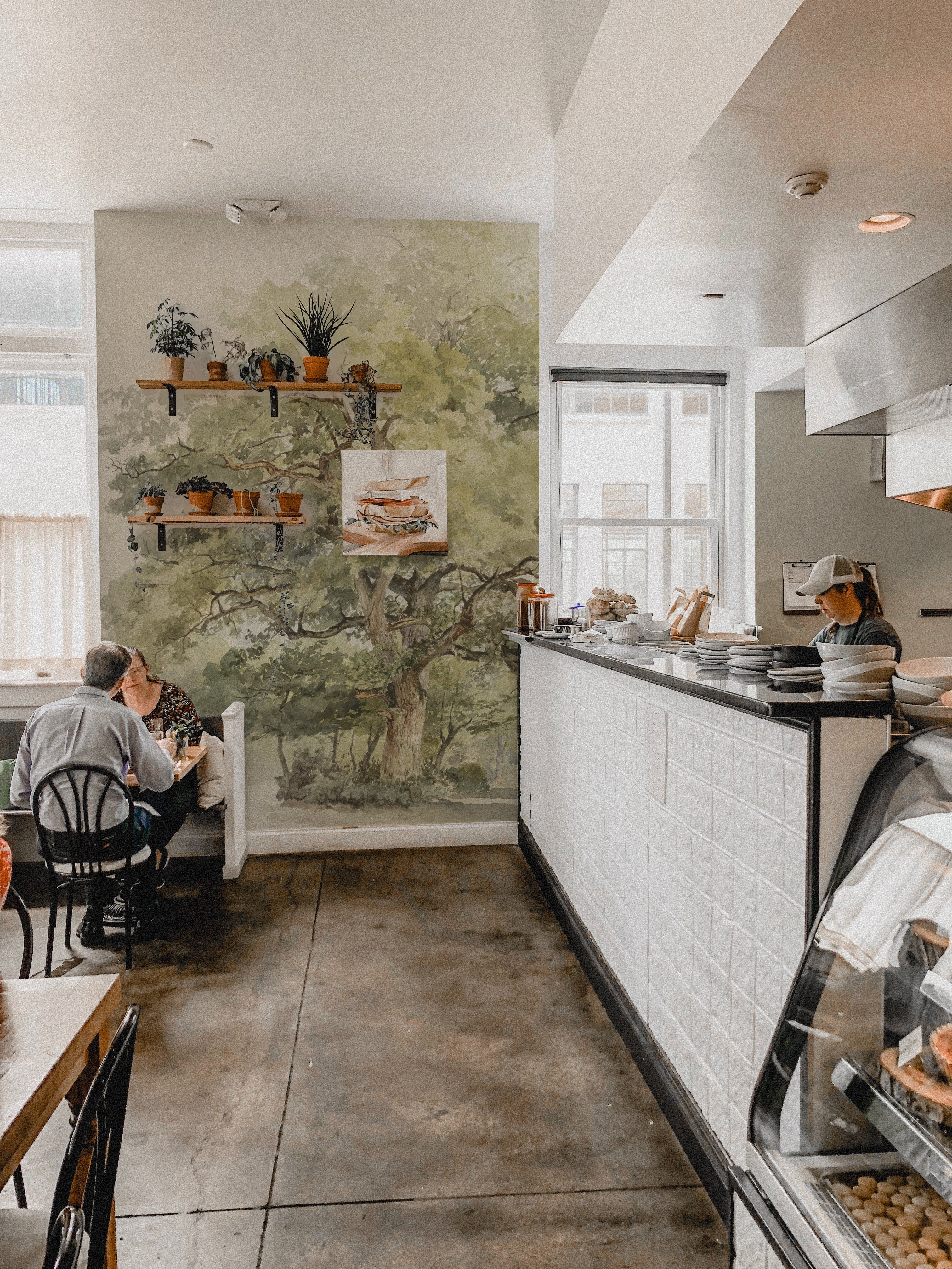 A cozy café interior where the Boomstudie Vintage Wall Mural serves as a backdrop, adding a rustic charm with its detailed depiction of a grand old tree. The mural provides a calming natural element to the space, harmonizing with wooden shelving filled with plants and cafe amenities.