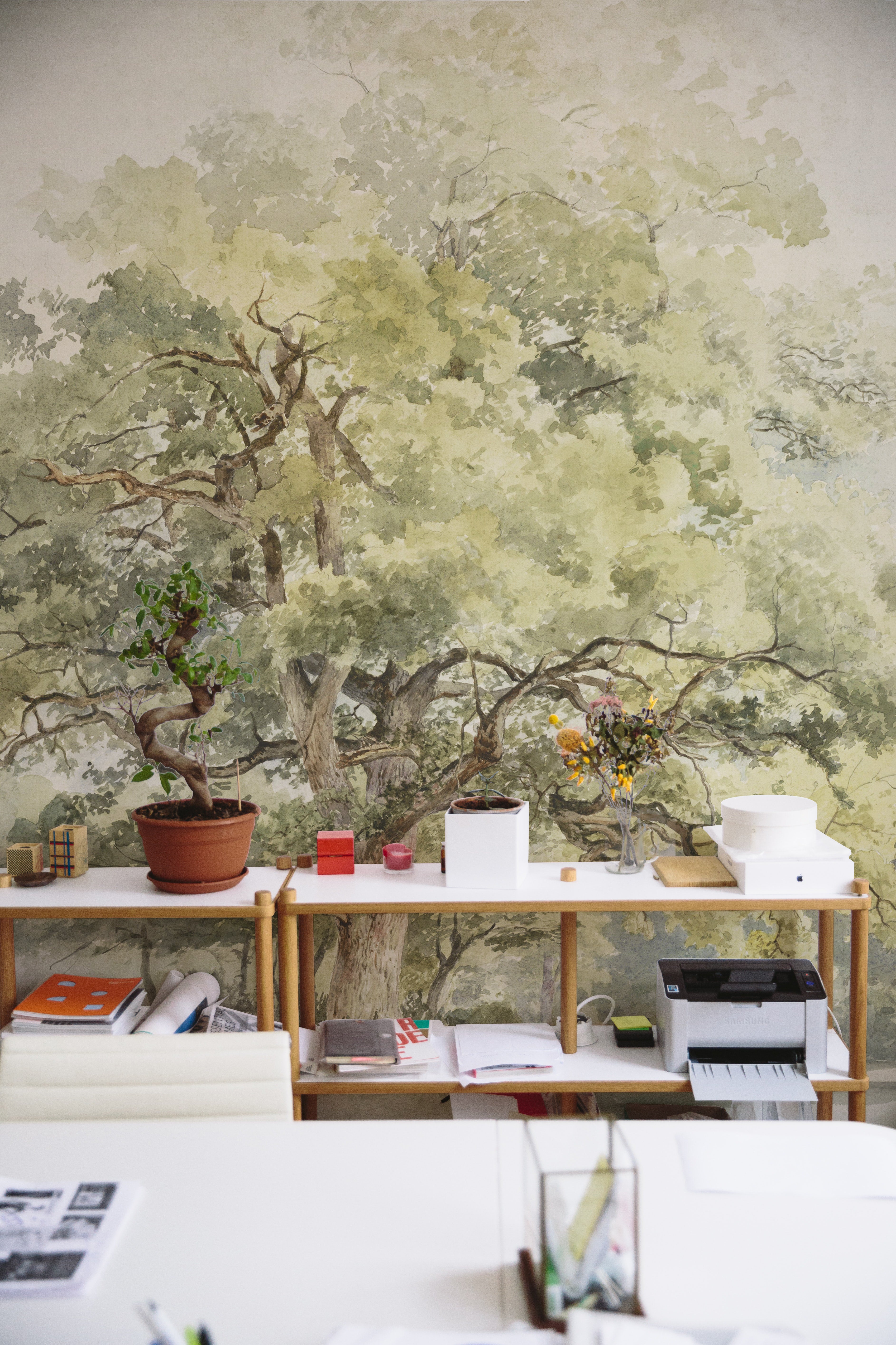 An office space where the Boomstudie Vintage Wall Mural creates a peaceful work environment. The mural, illustrating a dense tree with intricate branches and leaves, spans the entire wall, pairing beautifully with simple office furniture and decorative items to inspire creativity.