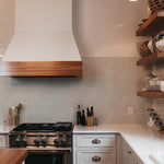 A stylish kitchen setting with 'William Bough Wallpaper' providing a soft backdrop to wooden open shelves, a white range hood, and stainless steel stove.