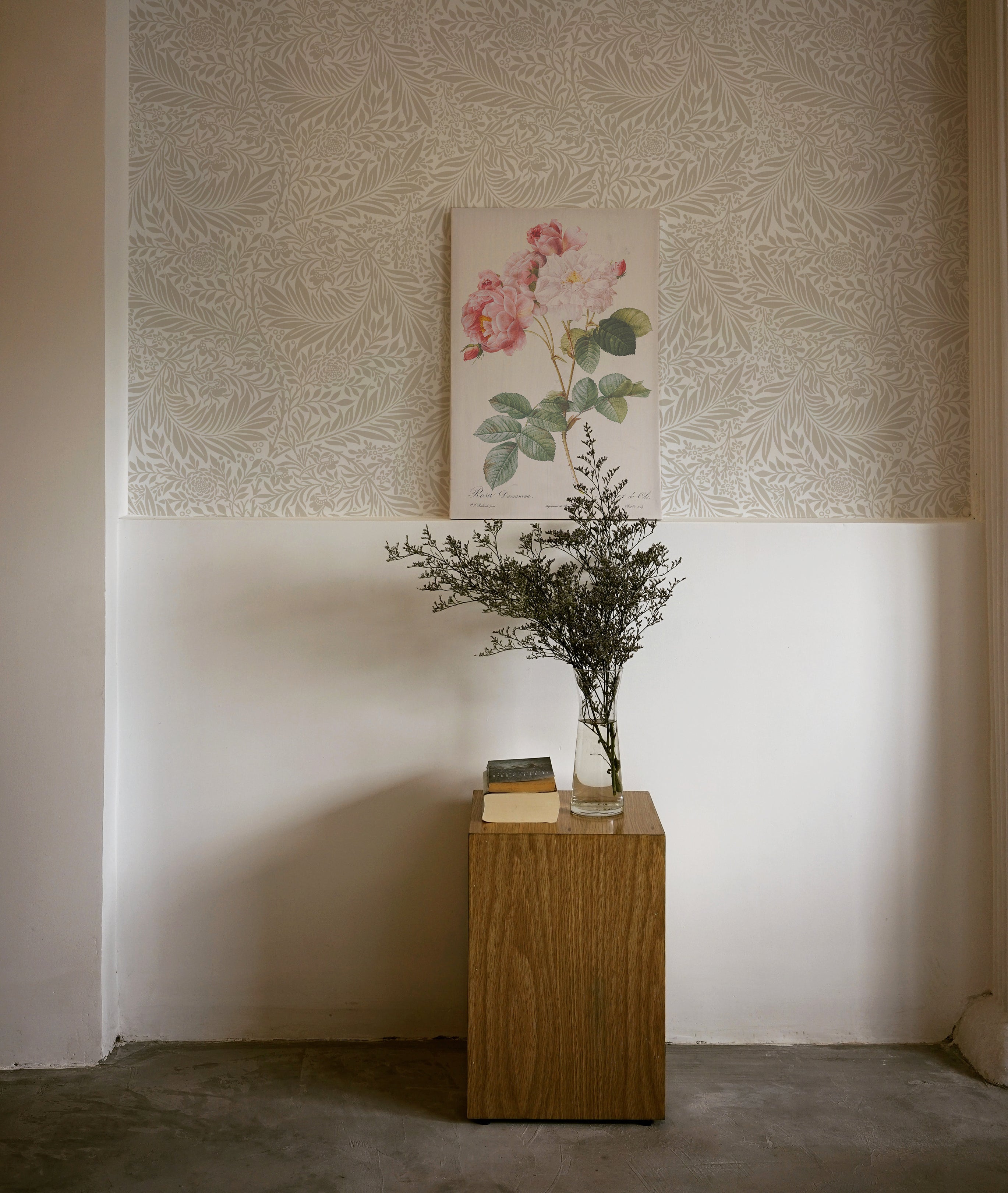 An interior corner showcasing the 'William Bough Wallpaper' with a large-leafed botanical pattern in muted tones, accented by a framed floral artwork and a rustic wooden pedestal with a vase and book.