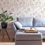 A contemporary living room featuring the "Atelier Botanique Wallpaper" with a minimalist botanical pattern in shades of grey. The elegant design provides a subtle backdrop to a modern grey sofa, accentuated by a black side table and a vibrant green houseplant, creating a serene and stylish space.