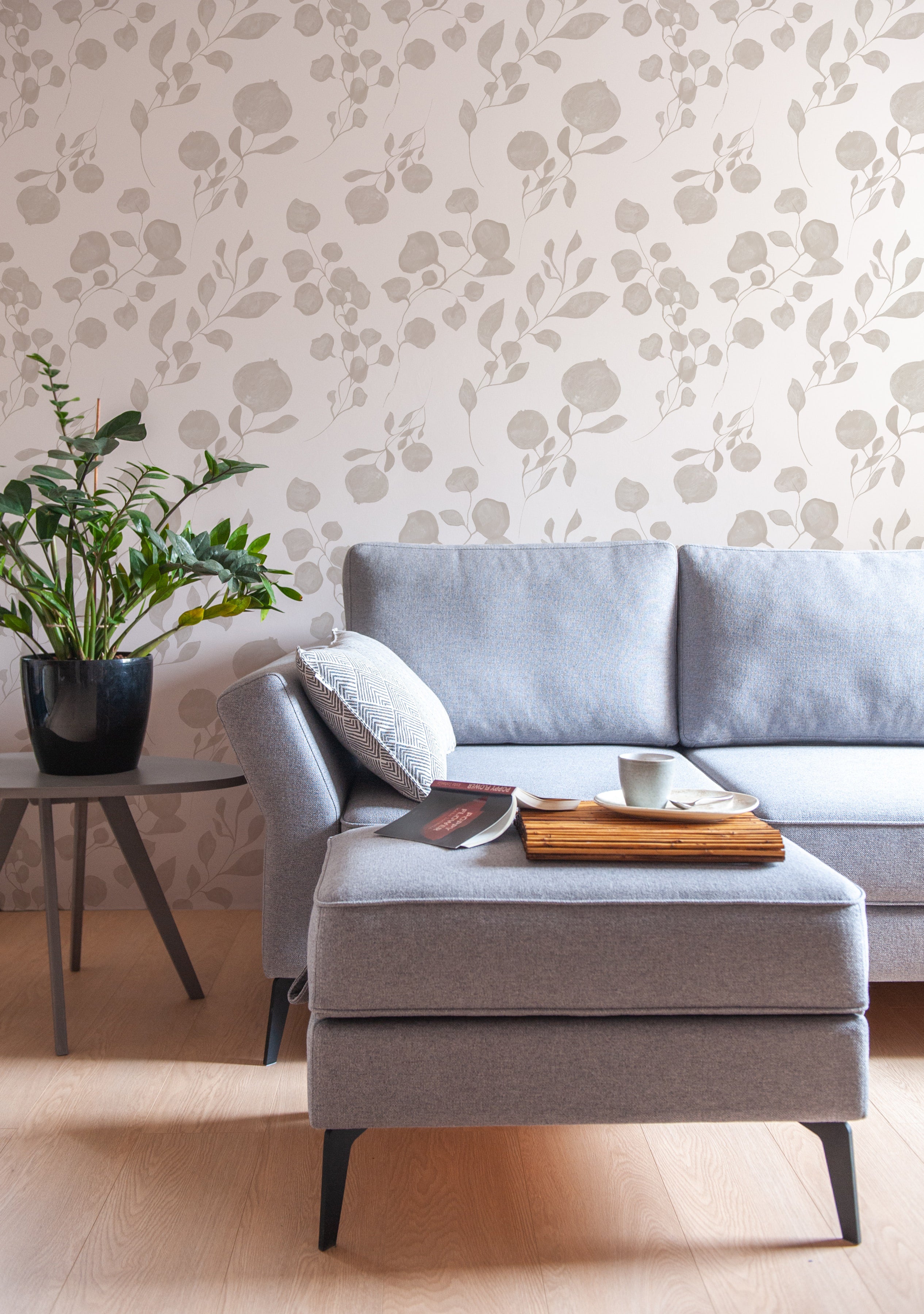 A contemporary living room featuring the "Atelier Botanique Wallpaper" with a minimalist botanical pattern in shades of grey. The elegant design provides a subtle backdrop to a modern grey sofa, accentuated by a black side table and a vibrant green houseplant, creating a serene and stylish space.