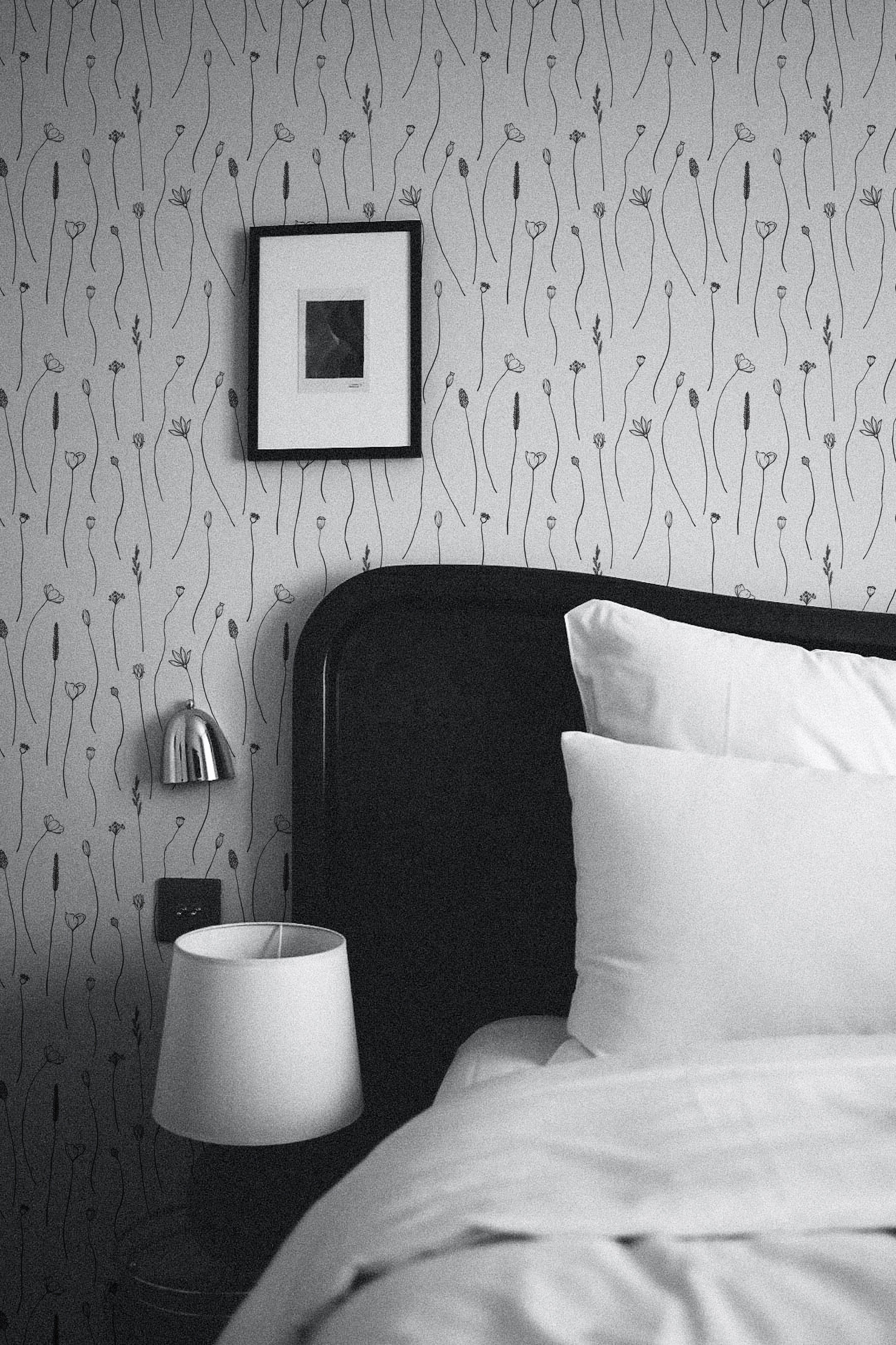 A stylish bedroom featuring the 'Wildflower Sketch Wallpaper' as a main decor element behind a classic black headboard. The wallpaper's subtle floral patterns add a touch of elegance and tranquility to the room, harmonizing with the simple and modern furnishings.