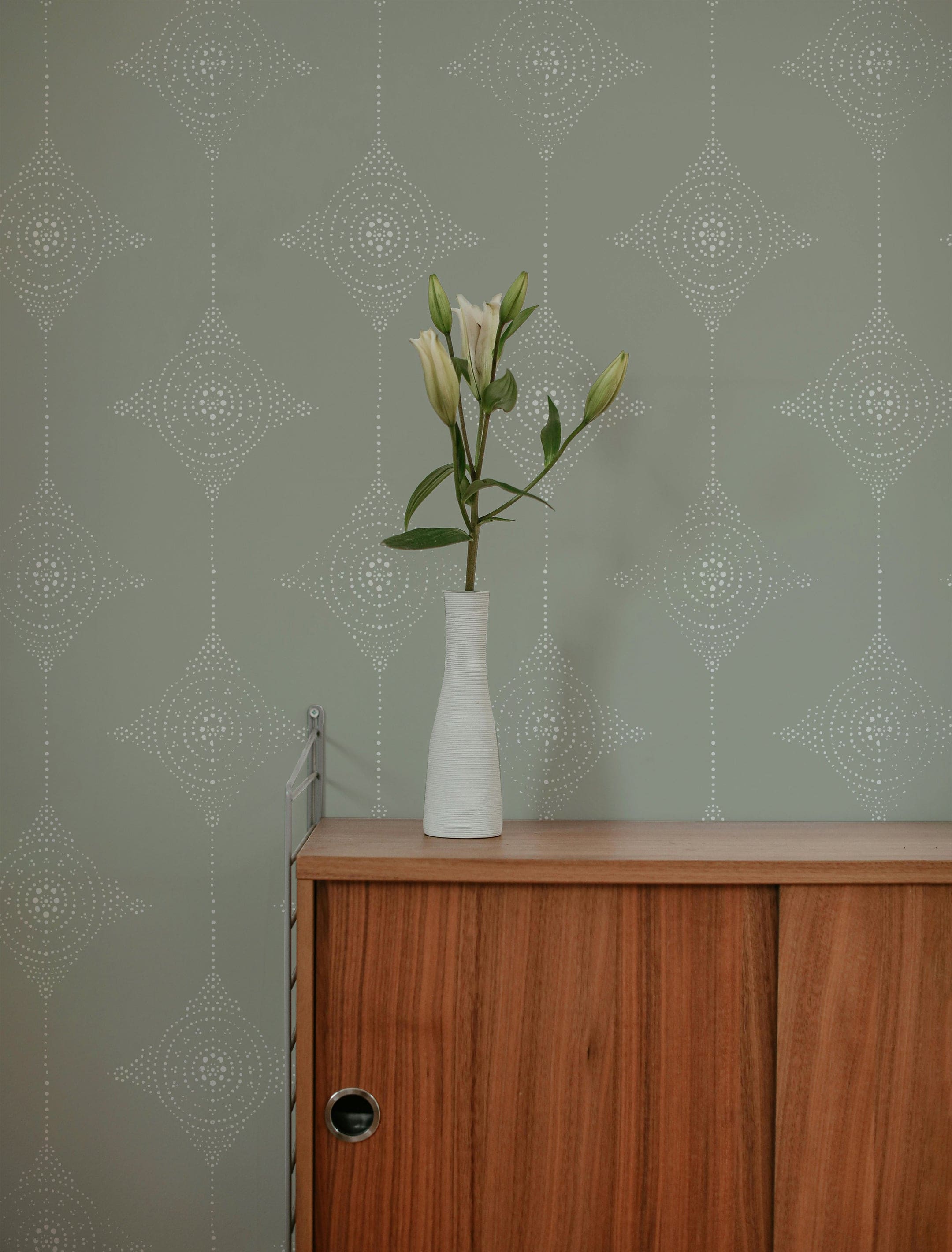 An interior scene featuring the Serenity Circles Wallpaper. The wallpaper's pattern of white circles on an olive green background provides a serene backdrop for a minimalist wooden console table adorned with a single white vase holding a blooming flower.