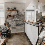 A cafe interior adorned with 'Kasteel Soelen - Wall Mural Wallpaper', featuring a castle surrounded by trees. The vintage-inspired mural creates a cozy and inviting atmosphere for the diners seated nearby.