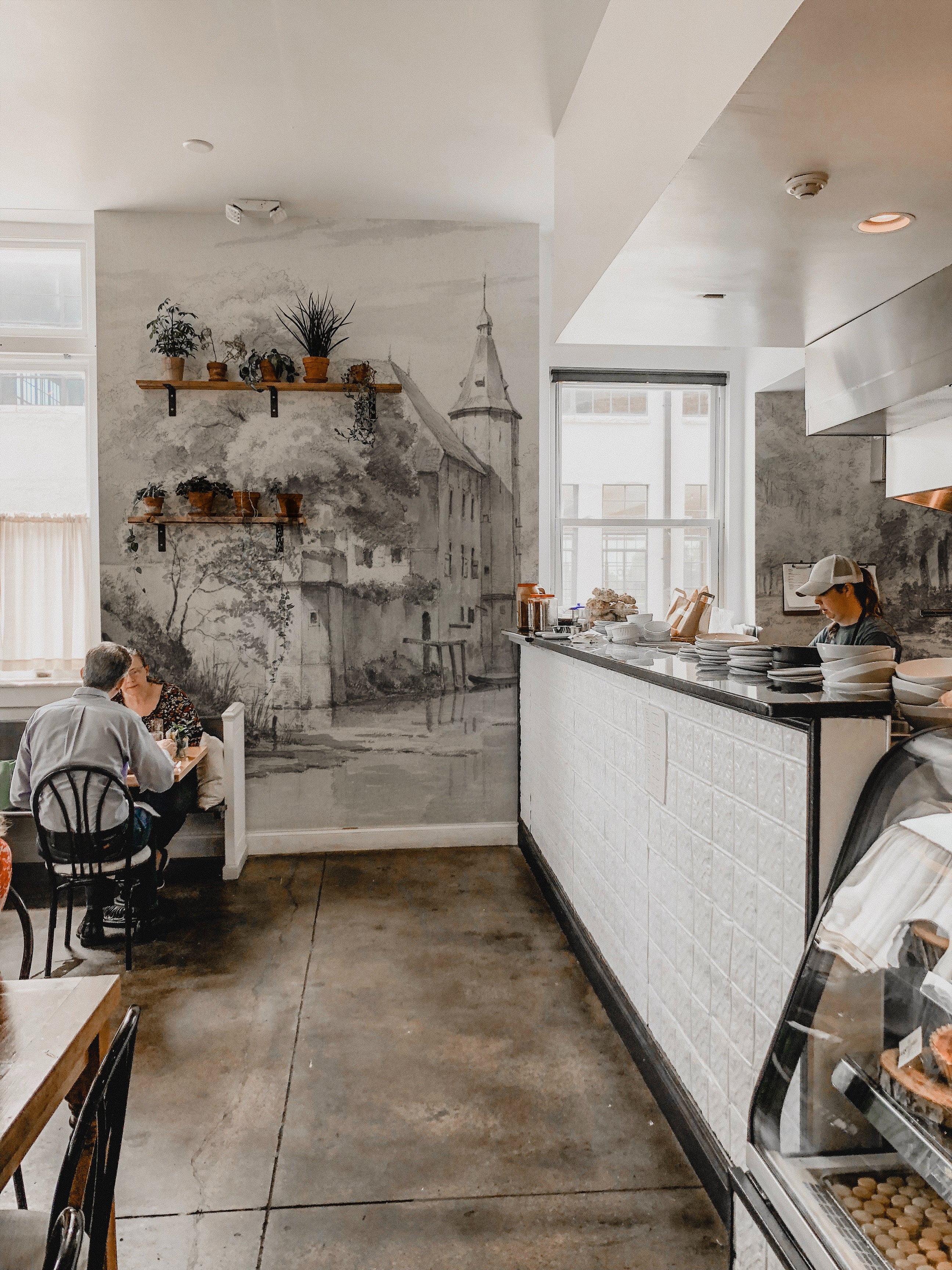 A cafe interior adorned with 'Kasteel Soelen - Wall Mural Wallpaper', featuring a castle surrounded by trees. The vintage-inspired mural creates a cozy and inviting atmosphere for the diners seated nearby.