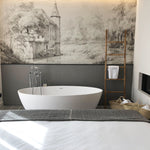Elegant bedroom design with the 'Kasteel Soelen - Wall Mural Wallpaper' as a backdrop behind a freestanding bathtub. The greyscale mural portrays a serene castle scene, adding a dramatic yet peaceful ambiance to the room