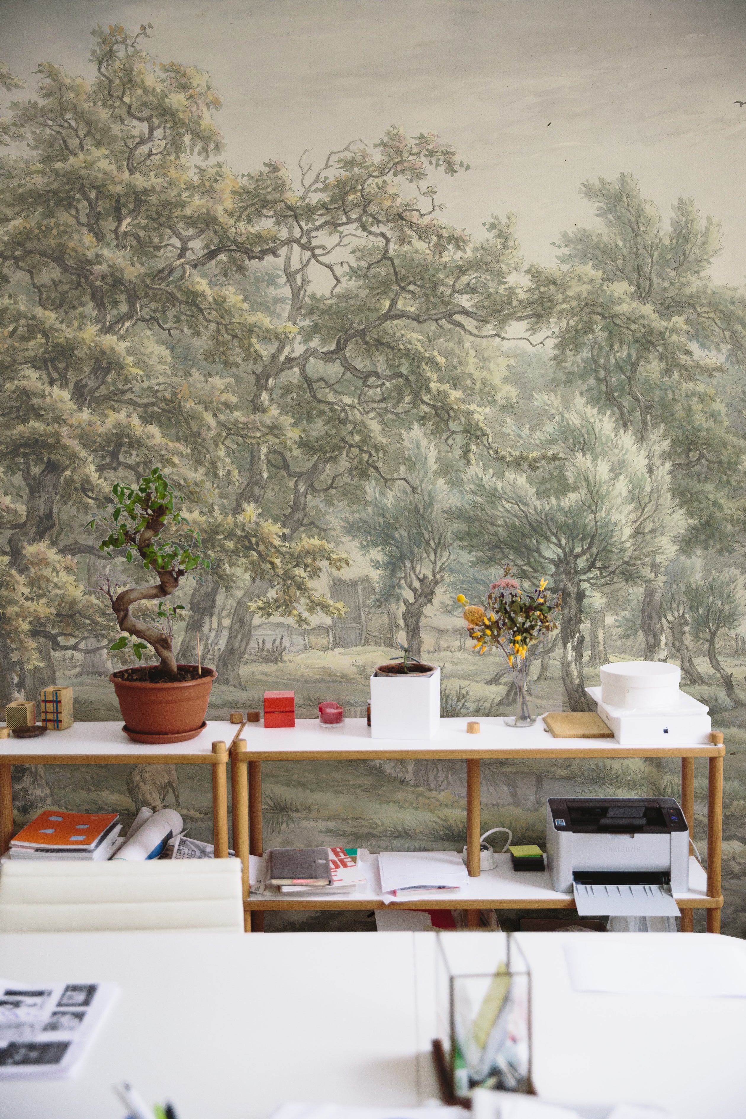 A vibrant workspace with a long wooden desk cluttered with office supplies, facing a wall mural depicting a serene 18th-century landscape scene from Eext with detailed trees and a pastoral setting.