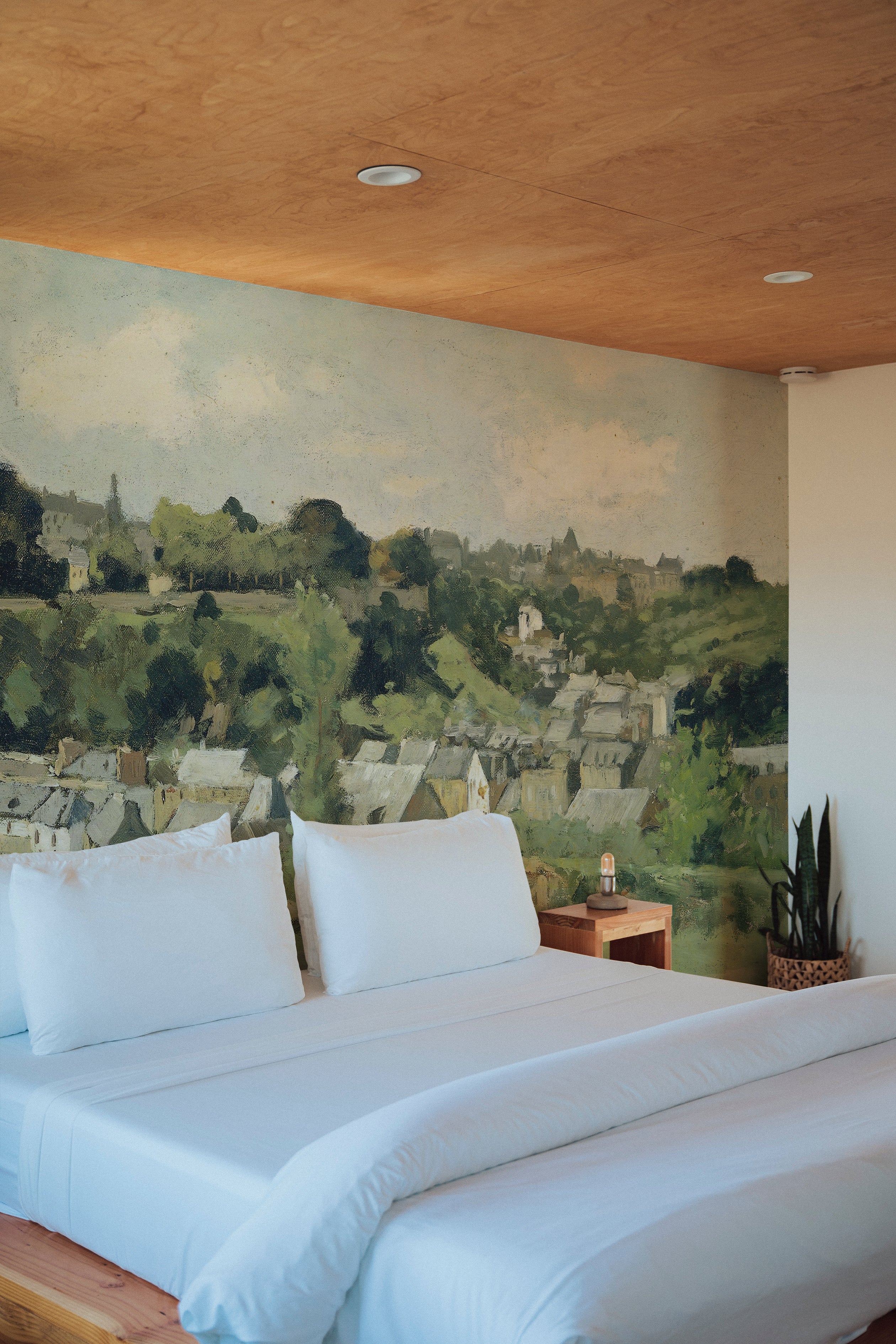 A bedroom setting enriched with a 'Views - Vintage Wall Mural', presenting a serene village scene. The mural serves as a stunning backdrop to a simple bed setup, complementing the room with its rich, earthy tones and peaceful landscape.