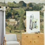 A vintage wall mural in a modern living setting, showcasing a pastoral landscape painting of a village. The artwork captures a soft, natural palette, offering a window into a tranquil rural life. A contemporary grey armchair and a minimalist wooden credenza accentuate the timeless charm of the mural.