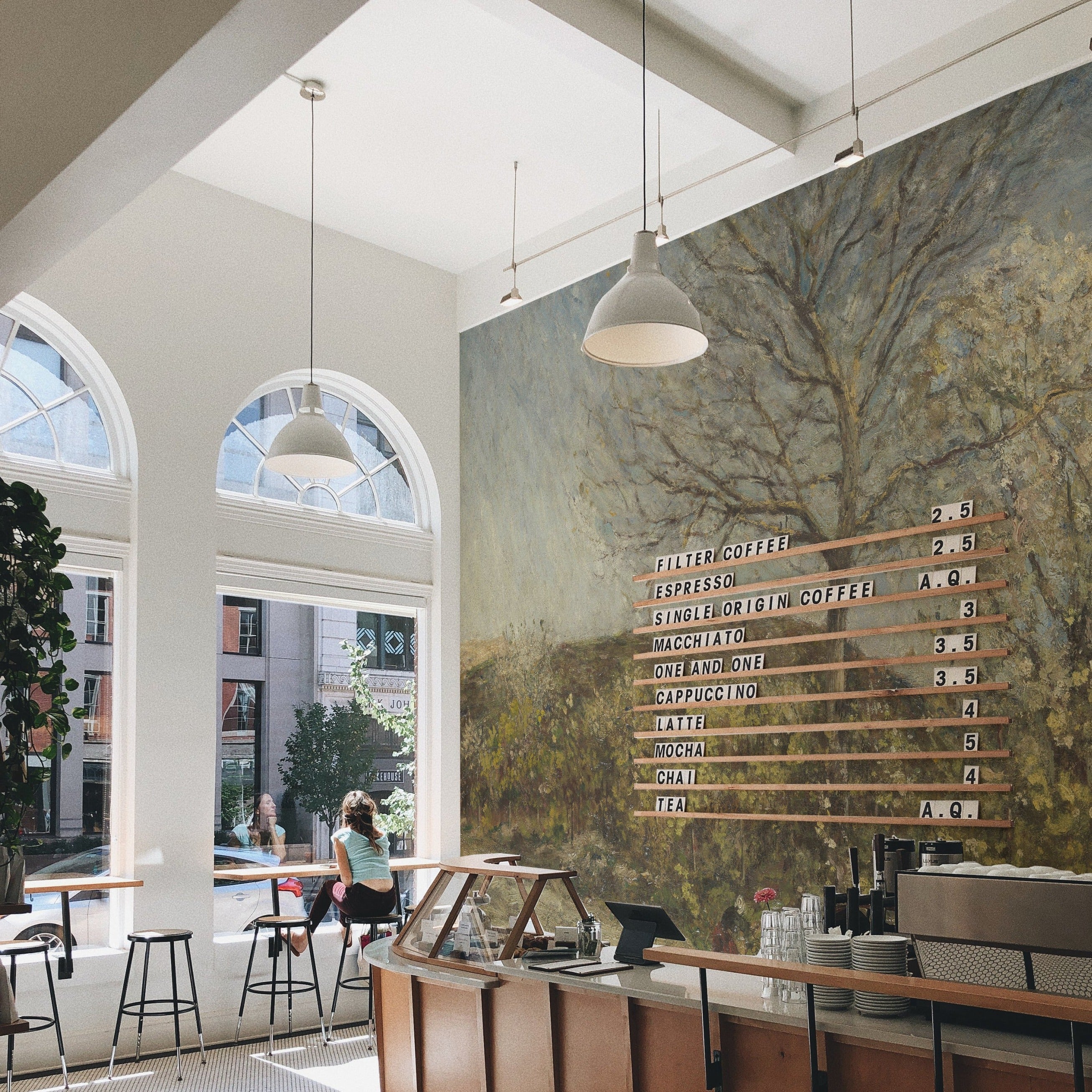 Coffee shop interior with a detailed landscape mural covering a large wall. The mural features a tree and vibrant foliage, adding a rustic charm to the modern cafe atmosphere, complete with wooden furniture and bright natural lighting.