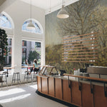 Coffee shop interior with a detailed landscape mural covering a large wall. The mural features a tree and vibrant foliage, adding a rustic charm to the modern cafe atmosphere, complete with wooden furniture and bright natural lighting.