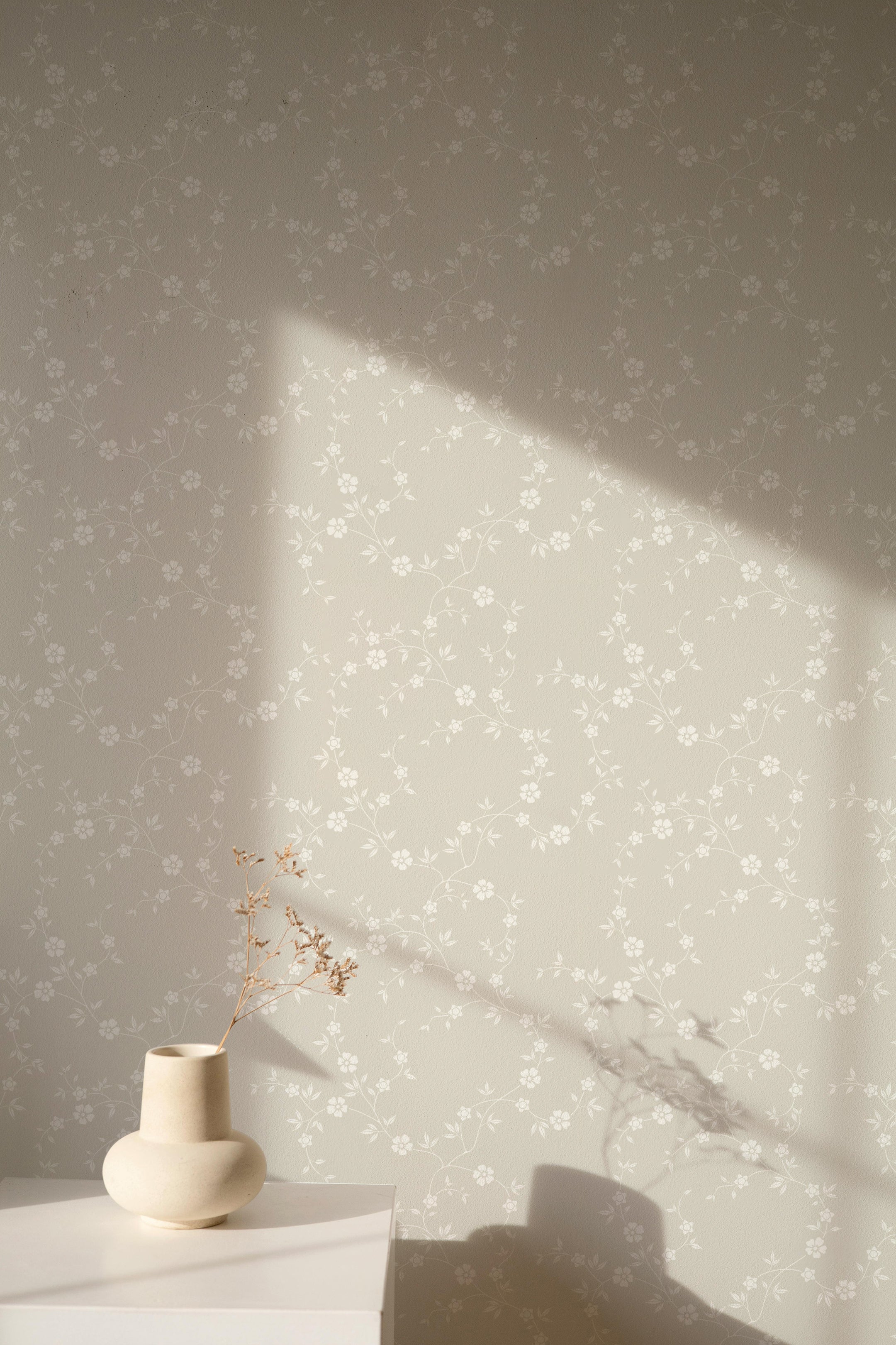 A minimalist interior scene featuring the Charming Floral Wallpaper II in a light beige background. The wallpaper's delicate white floral pattern is complemented by a small white table and a simple ceramic vase with dried flowers, creating a peaceful and elegant setting.