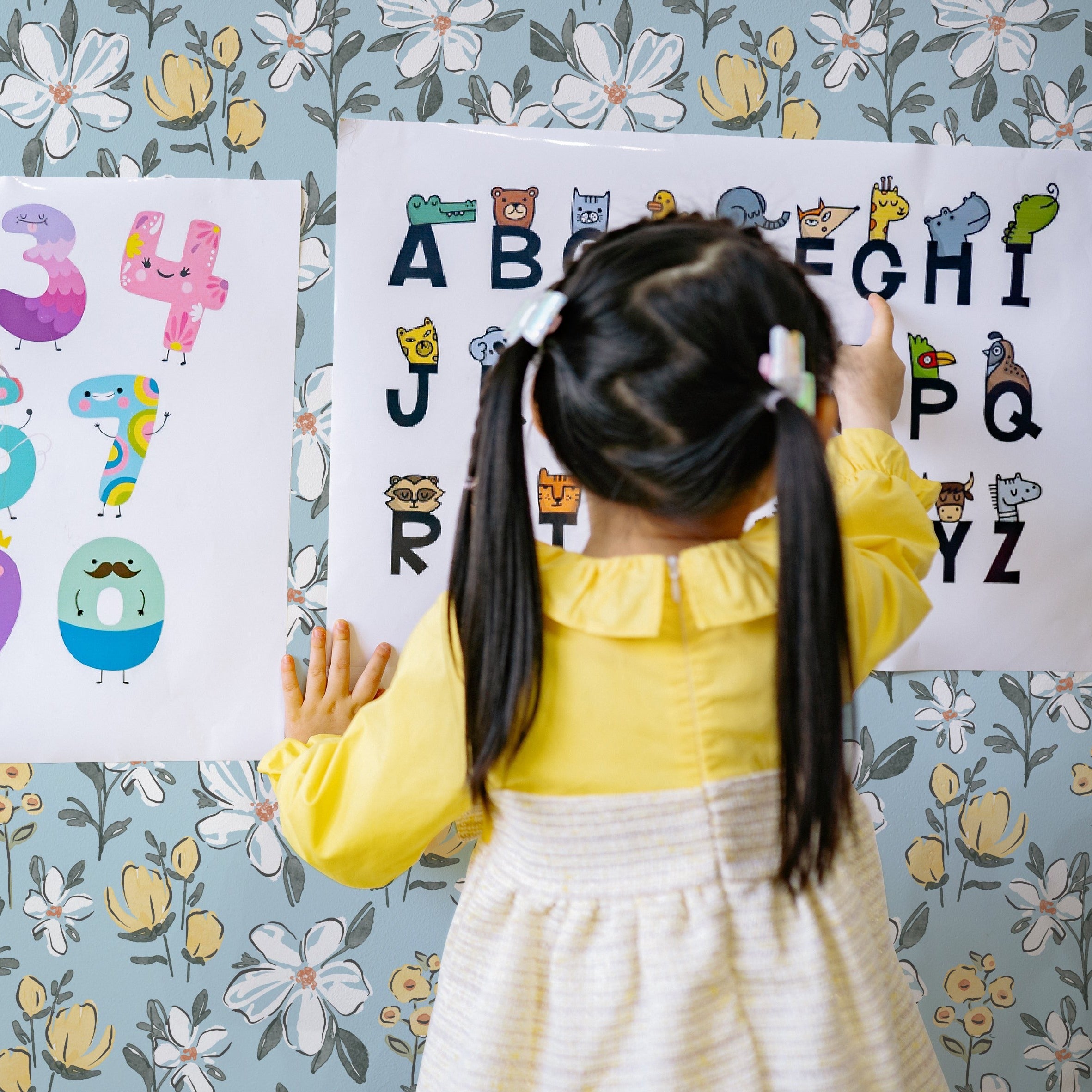 A young child in a yellow dress is interacting with an educational alphabet poster against a backdrop of the Sweet Nursery Wallpaper, enhancing the playful and educational atmosphere of the room.