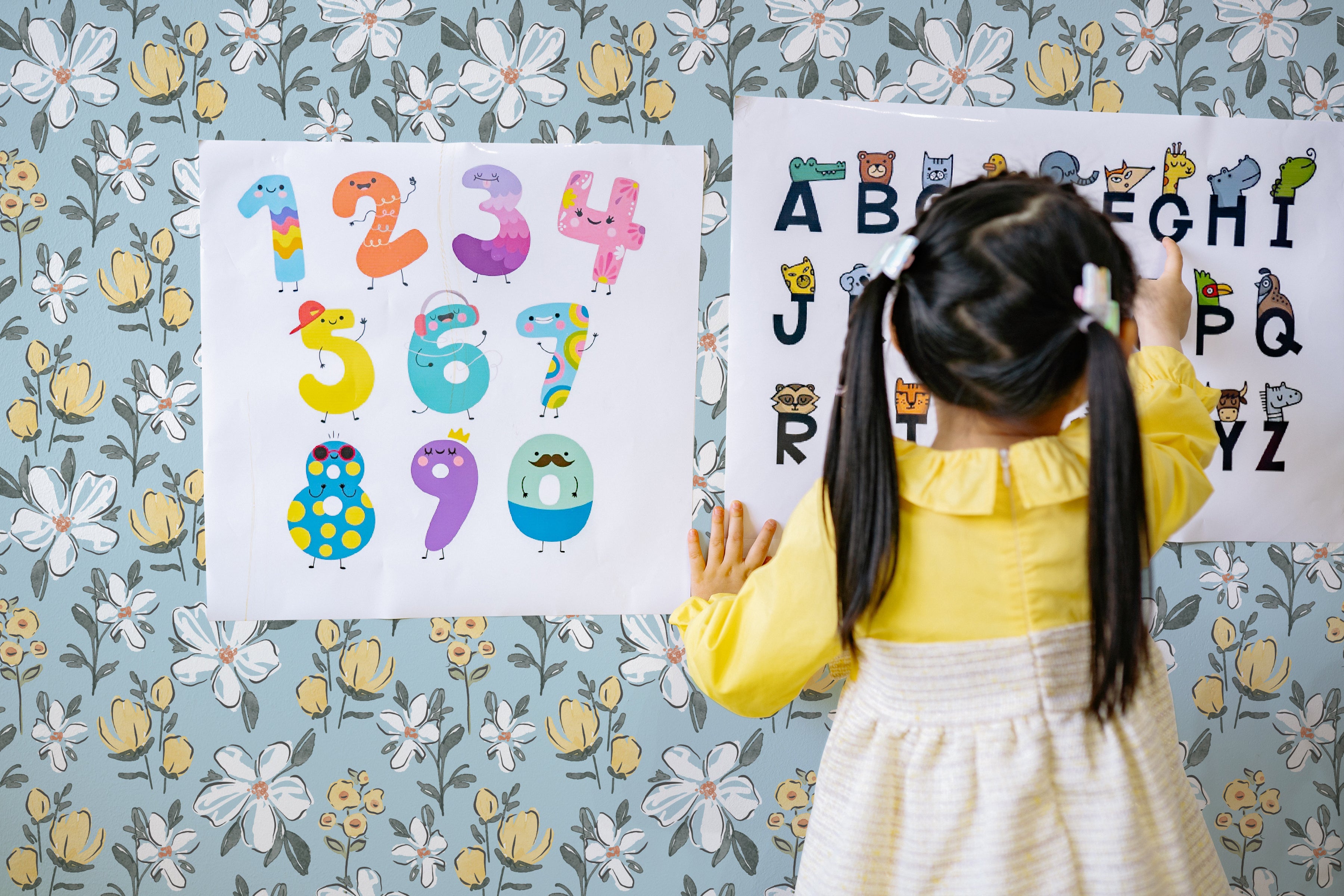 A young child in a yellow dress is interacting with an educational alphabet poster against a backdrop of the Sweet Nursery Wallpaper, enhancing the playful and educational atmosphere of the room.