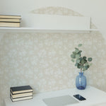 A minimalist home office setup with a white desk and chair against a wall covered with Vintage Garden Floral Wallpaper in ecru, showcasing a subtle floral pattern. The wallpaper imparts an elegant backdrop to the simple white shelf holding books, a blue vase with eucalyptus branches, a closed laptop, and a smartphone.