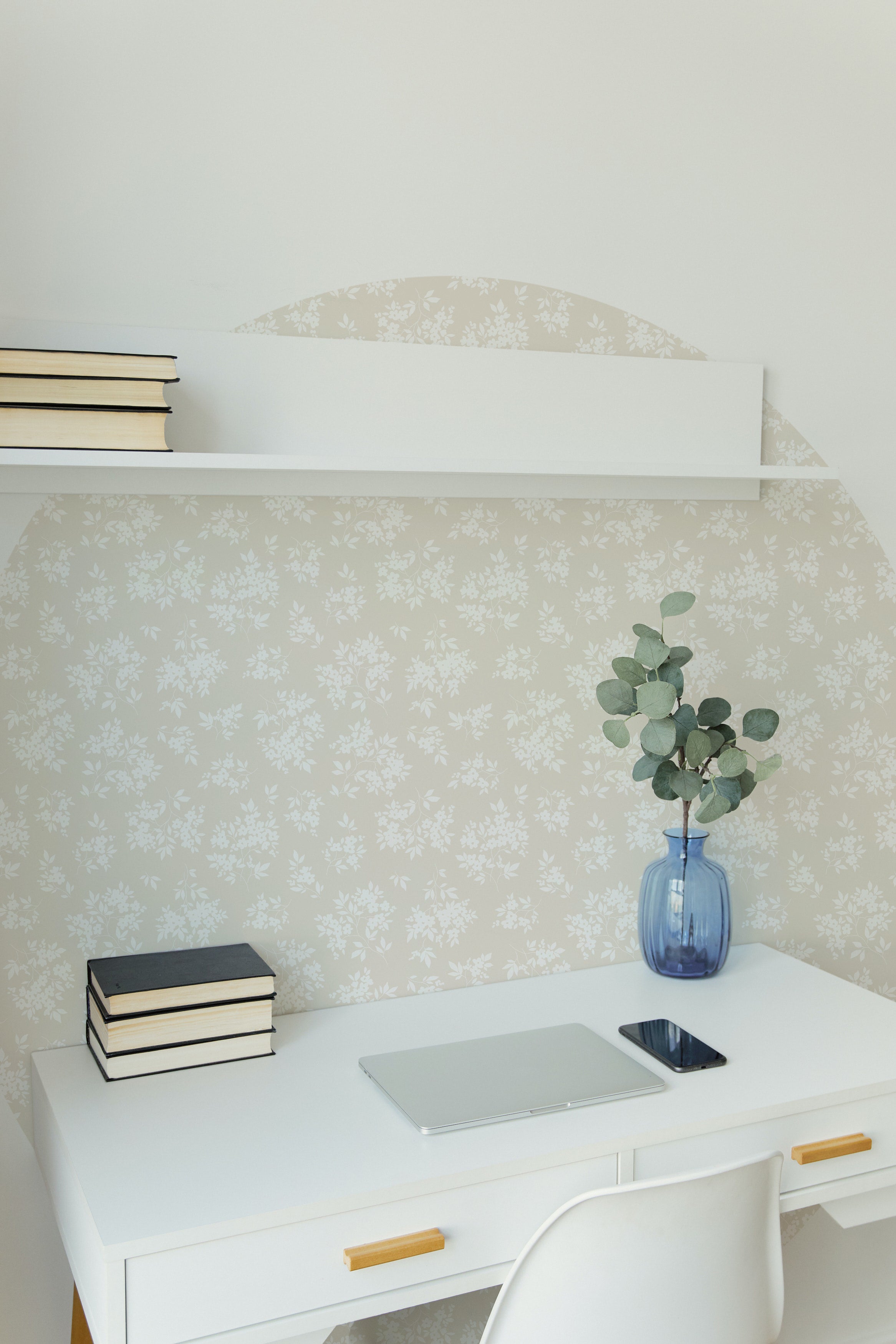 A minimalist home office setup with a white desk and chair against a wall covered with Vintage Garden Floral Wallpaper in ecru, showcasing a subtle floral pattern. The wallpaper imparts an elegant backdrop to the simple white shelf holding books, a blue vase with eucalyptus branches, a closed laptop, and a smartphone.
