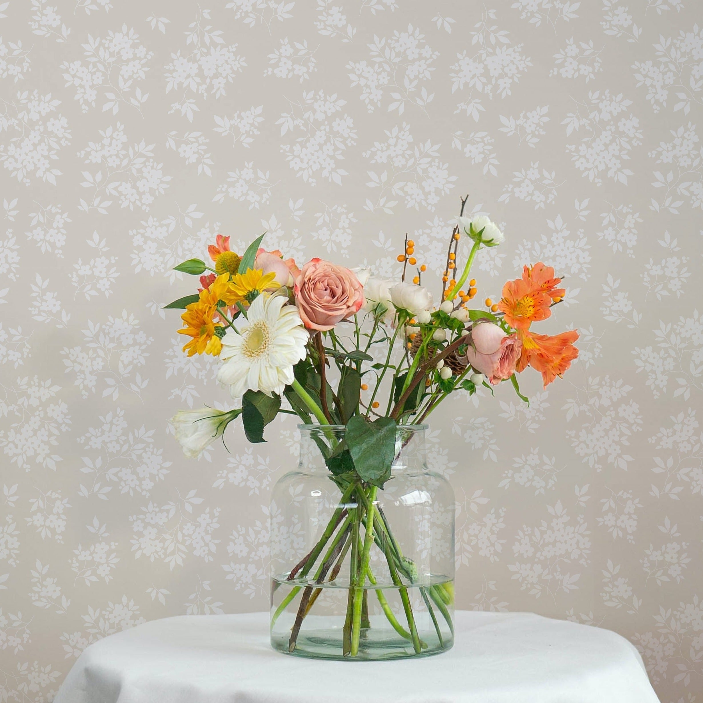 A vibrant bouquet of flowers in warm tones of orange, yellow, and pink, arranged in a clear glass vase on a table, with the Vintage Garden Floral Wallpaper - Ecru providing a subtle and sophisticated floral pattern in the backdrop, complementing the vivid colors of the bouquet.