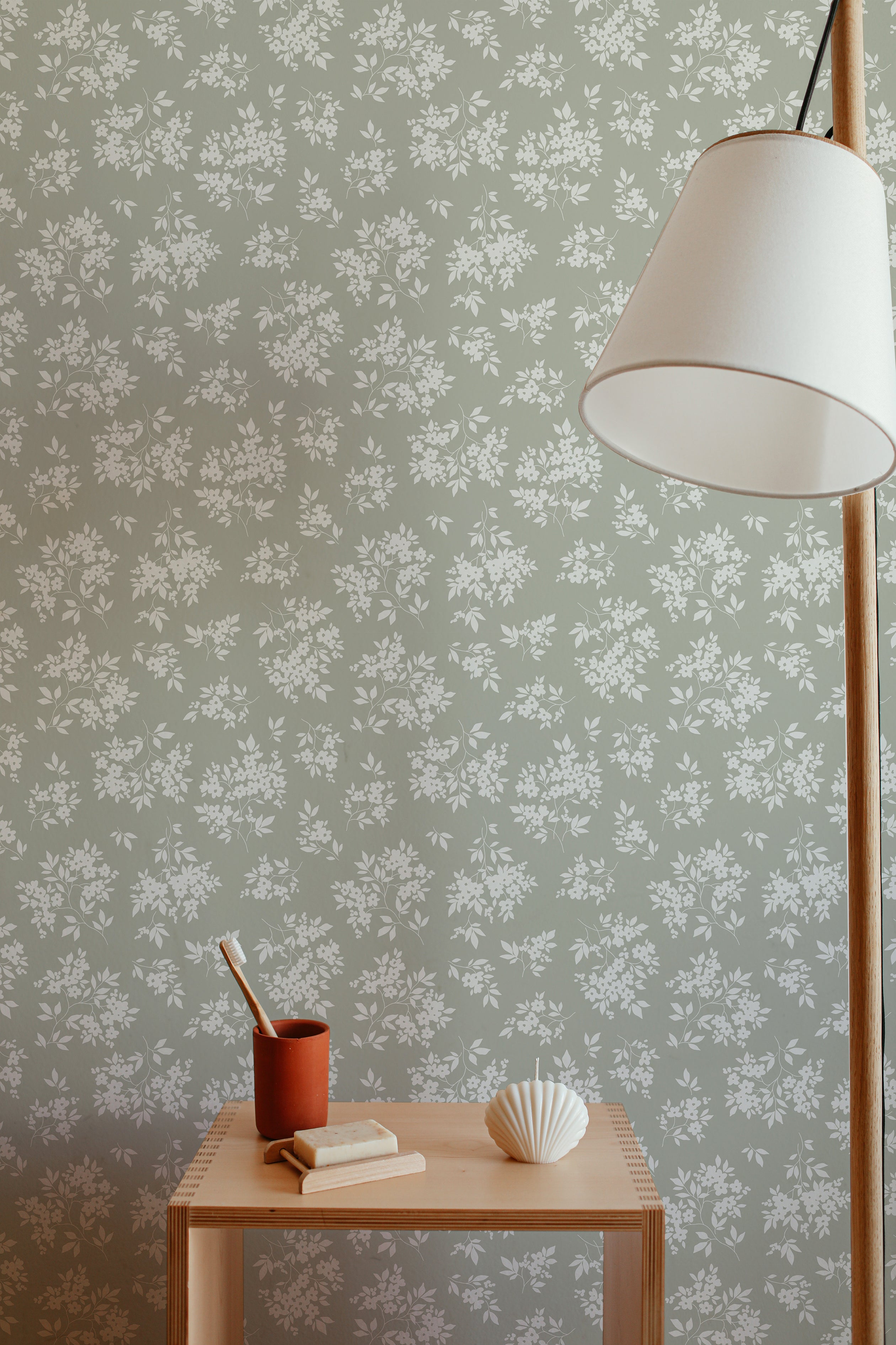 A cozy corner of a room featuring a minimalist wooden side table against a wall covered with Vintage Garden Floral Wallpaper in Olive. On the table, there's a terracotta mug holding a toothbrush, a bar of soap, and a decorative shell-shaped object, invoking a sense of tranquility and simplicity. A modern floor lamp with a white shade casts a soft glow on the scene.