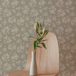 A simple, elegant wooden chair stands before a wall adorned with Vintage Garden Floral Wallpaper in Olive. On the chair, a white vase with budding green flowers adds a touch of life and color to the serene setting, suggesting a harmonious blend of modern design and nature-inspired elements.