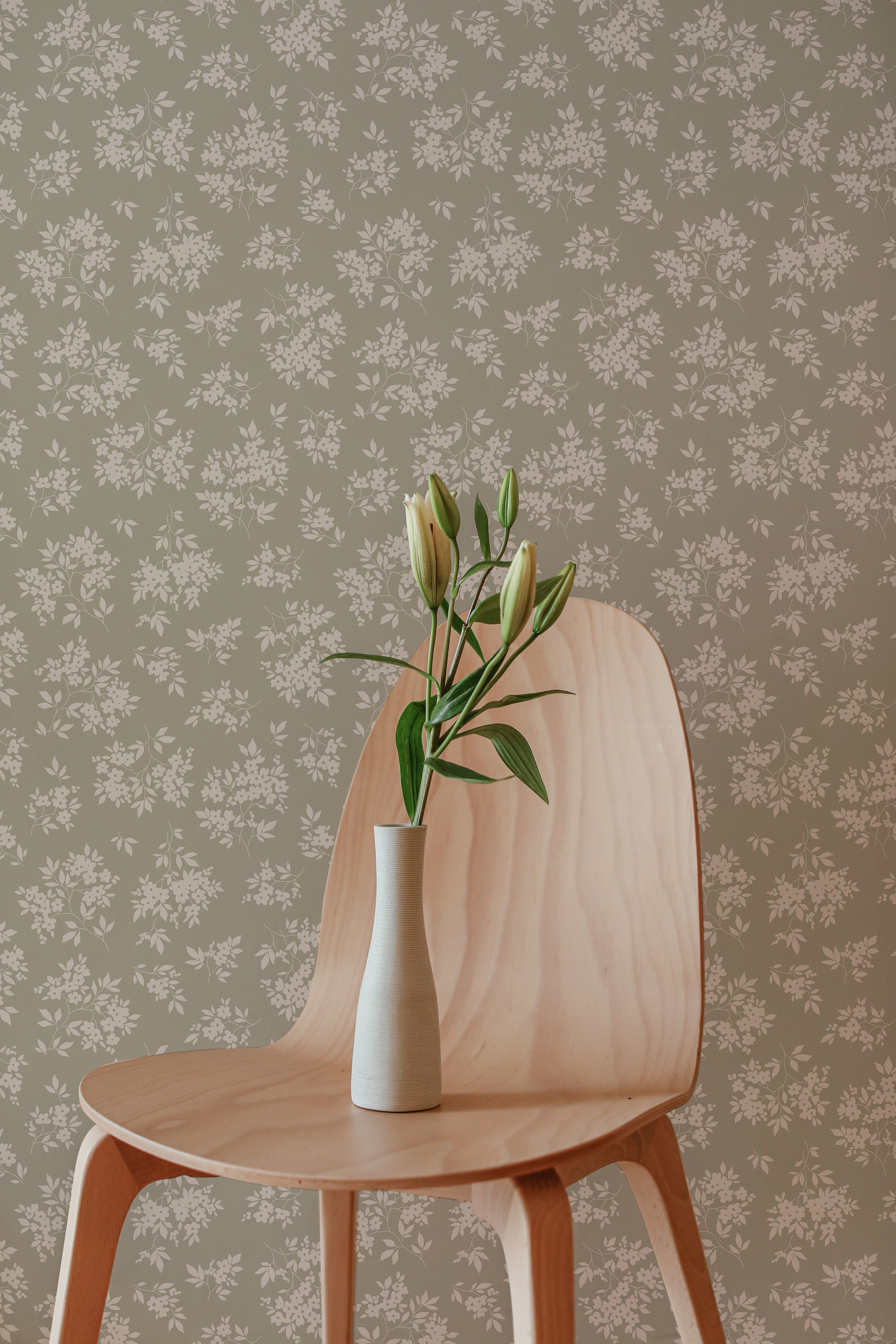 A simple, elegant wooden chair stands before a wall adorned with Vintage Garden Floral Wallpaper in Olive. On the chair, a white vase with budding green flowers adds a touch of life and color to the serene setting, suggesting a harmonious blend of modern design and nature-inspired elements.