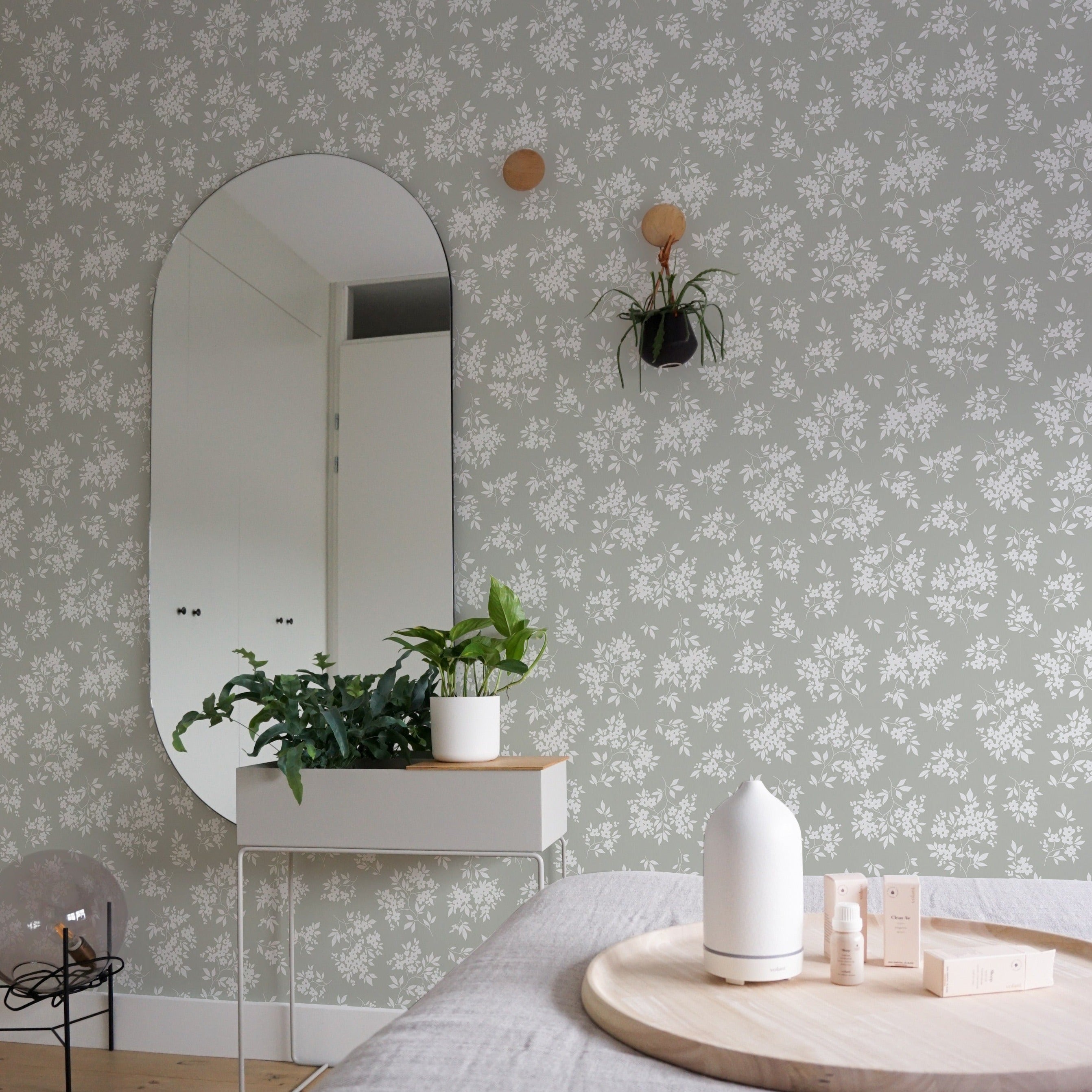 A tasteful interior featuring the Vintage Garden Floral Wallpaper in Olive as a backdrop. A sleek, oval-shaped mirror is centered on the wall, with a floating shelf below holding a lush potted plant and a white diffuser. To the right, hanging planters add vertical interest, completing the room's fresh and organic aesthetic.