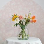 A charming still life of a vibrant floral arrangement in a clear glass vase, set against a delicate champagne-colored wallpaper adorned with white floral patterns, enhancing a soft, serene ambiance.