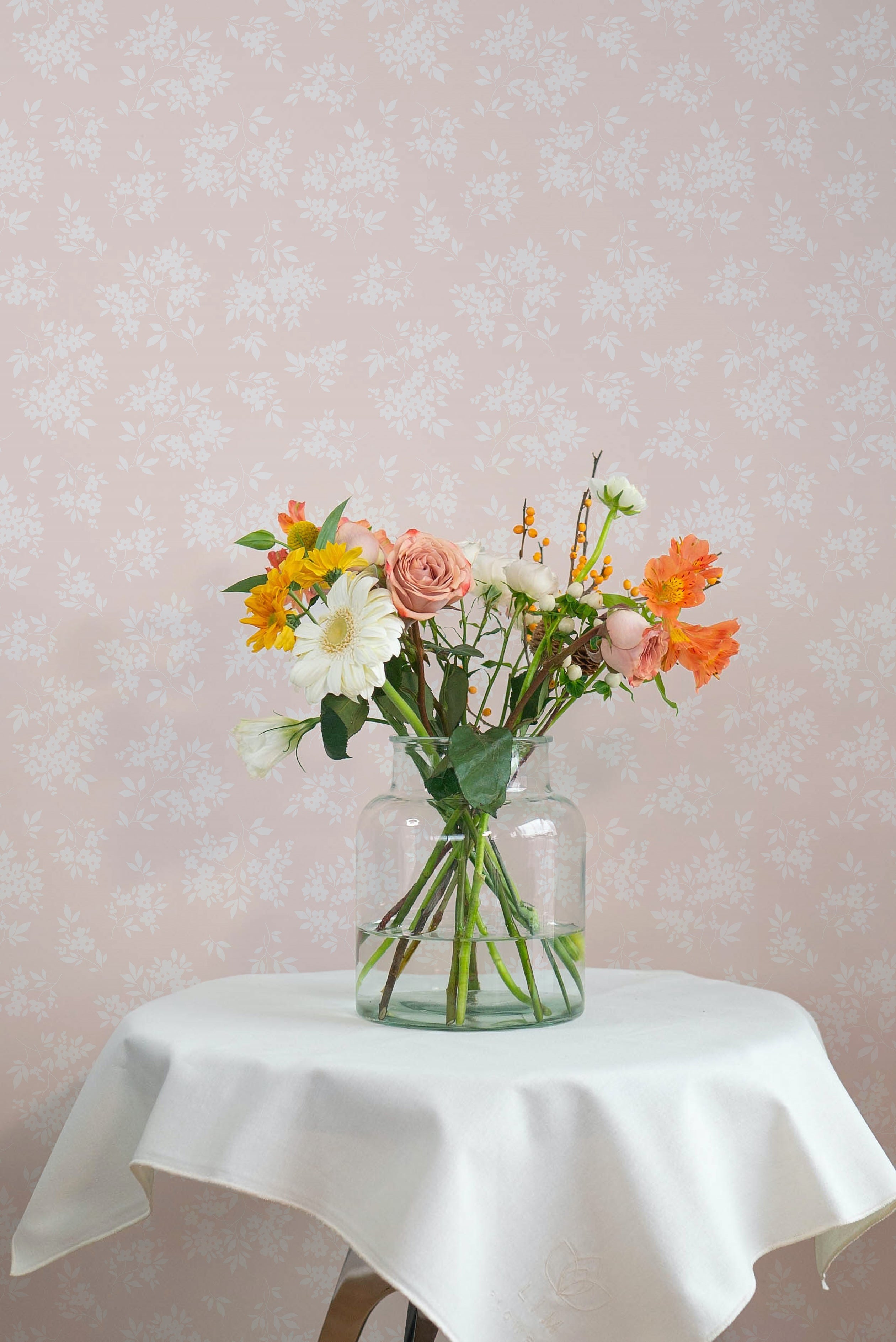 A charming still life of a vibrant floral arrangement in a clear glass vase, set against a delicate champagne-colored wallpaper adorned with white floral patterns, enhancing a soft, serene ambiance.