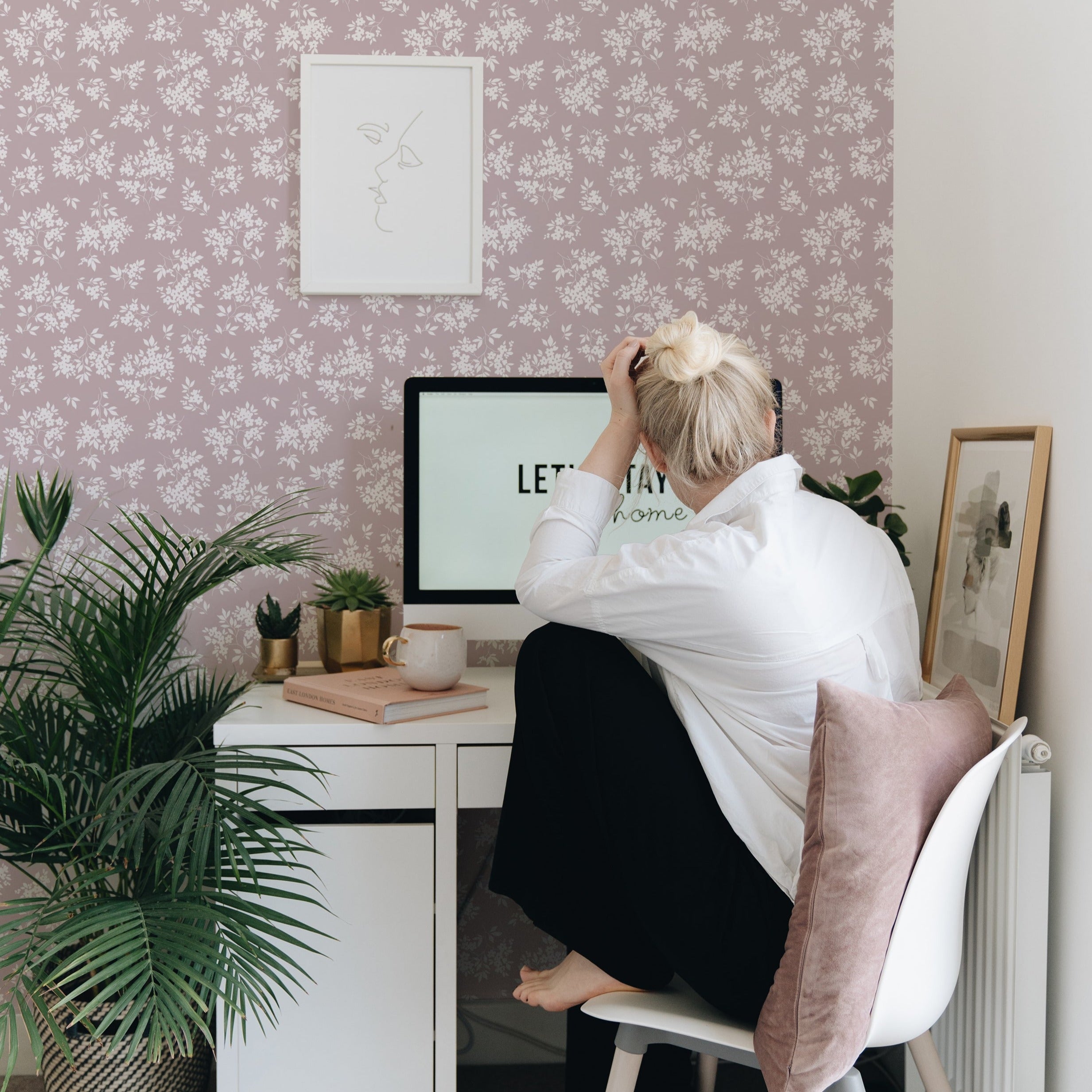 A home office setting where the Vintage Garden Floral Wallpaper is featured on a wall, providing a charming backdrop for a work environment. A person sits facing a computer with their back to the camera, creating an atmosphere of casual professionalism. The wallpaper adds a cozy and calming aesthetic to the space, complemented by indoor plants and minimalist desk accessories.
