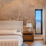 A cozy bedroom showcasing the "Parade" wallpaper mural, featuring a detailed cityscape. The room includes a plush bed, wooden side table, and a large window revealing a distant mountain view, blending indoor artistry with outdoor beauty.