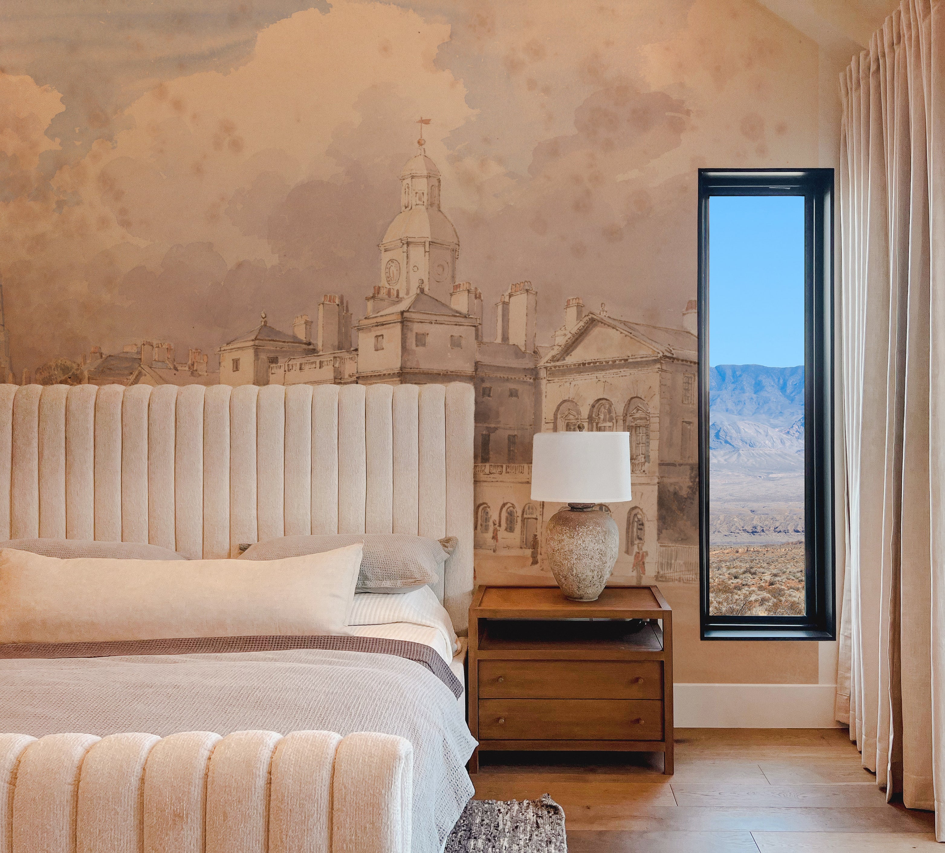 A cozy bedroom showcasing the "Parade" wallpaper mural, featuring a detailed cityscape. The room includes a plush bed, wooden side table, and a large window revealing a distant mountain view, blending indoor artistry with outdoor beauty.