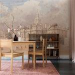 A sophisticated dining room displaying the "Parade" wallpaper mural depicting a historic cityscape with intricate architectural details. A wooden table, matching chairs, and a vase with pink blossoms enhance the room's warmth.