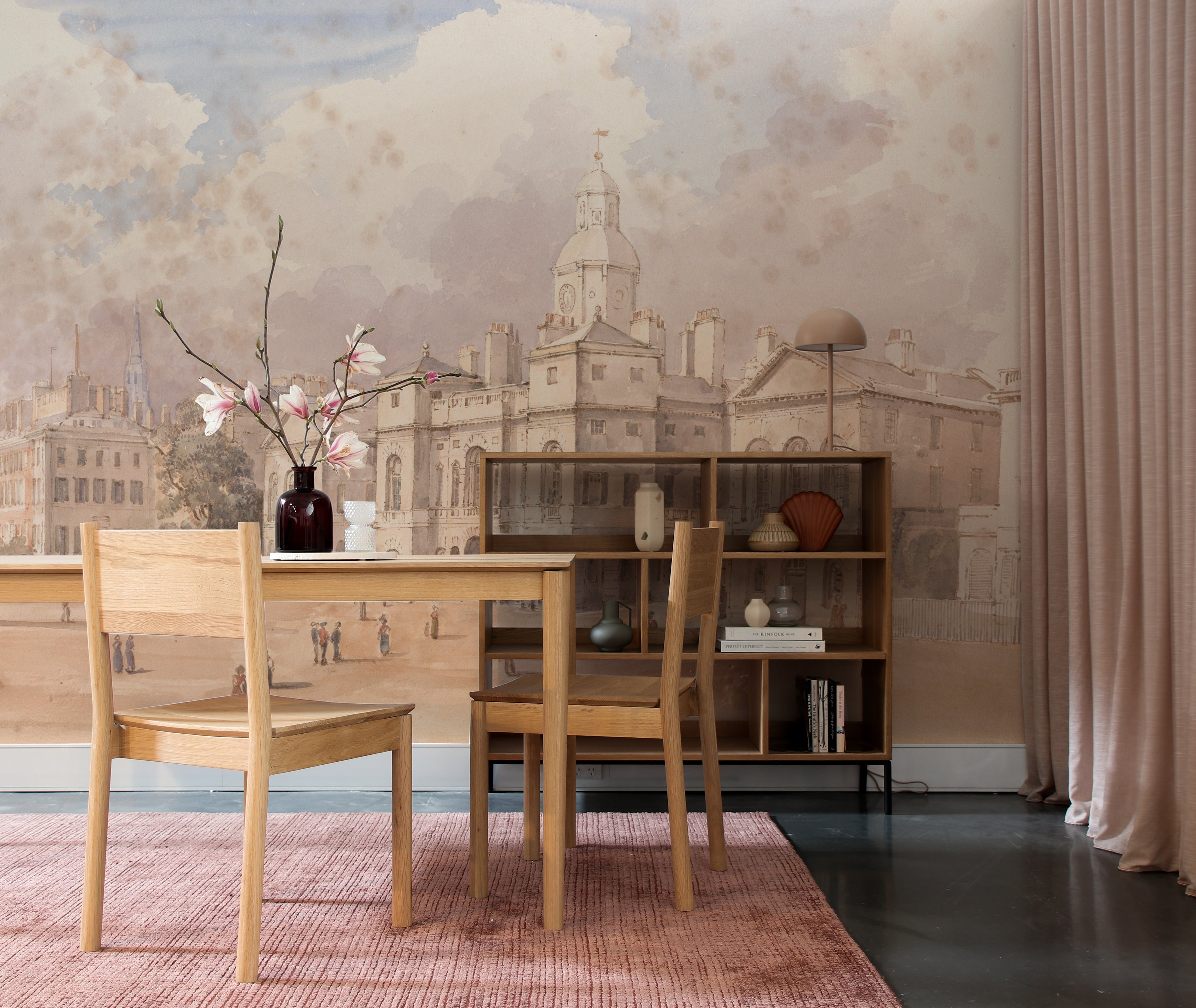 A sophisticated dining room displaying the "Parade" wallpaper mural depicting a historic cityscape with intricate architectural details. A wooden table, matching chairs, and a vase with pink blossoms enhance the room's warmth.