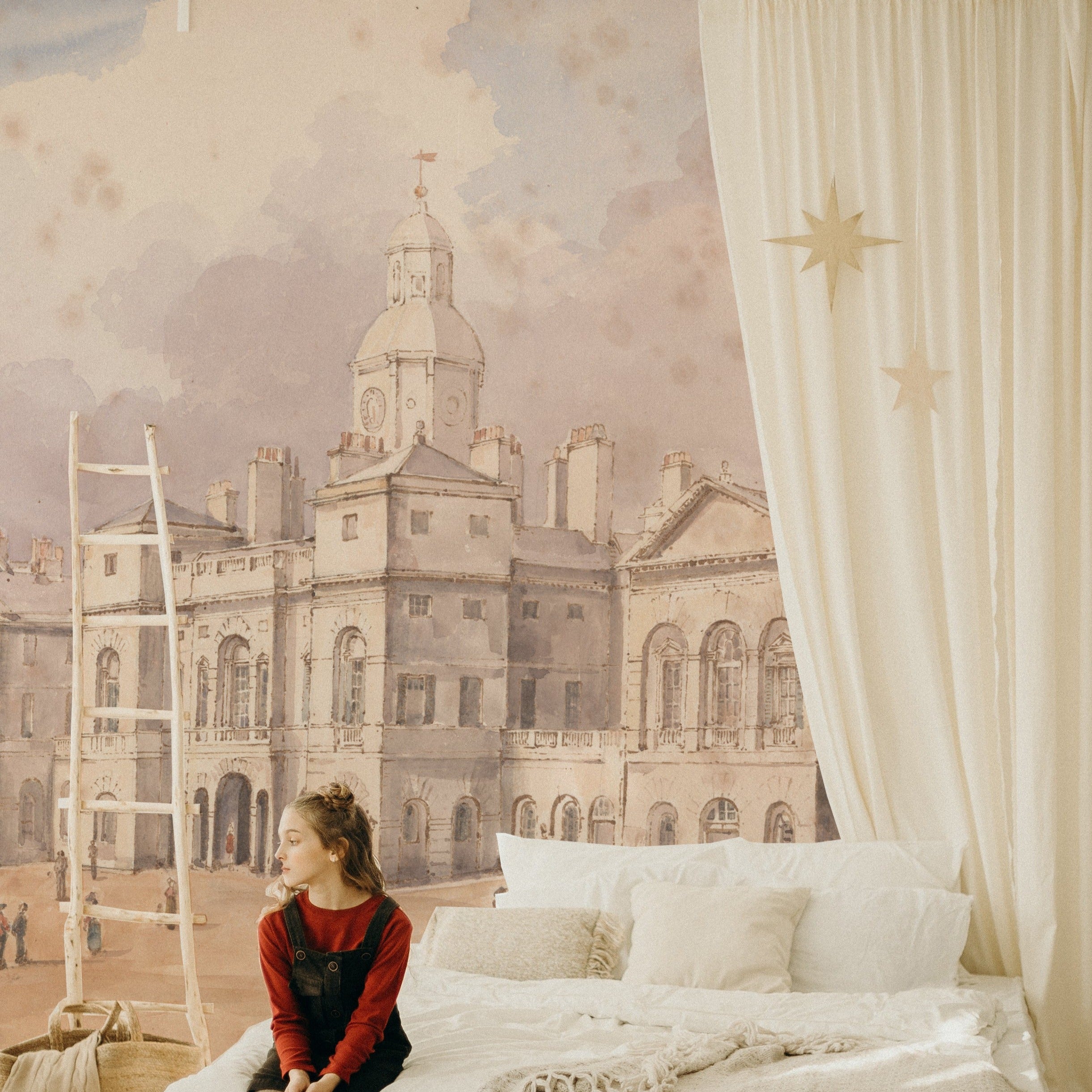 A child's bedroom with a whimsical touch, featuring the "Parade" wallpaper mural as a backdrop. The mural's historic cityscape is paired with simple, cozy bedding and a star-adorned curtain, creating a dreamy atmosphere.