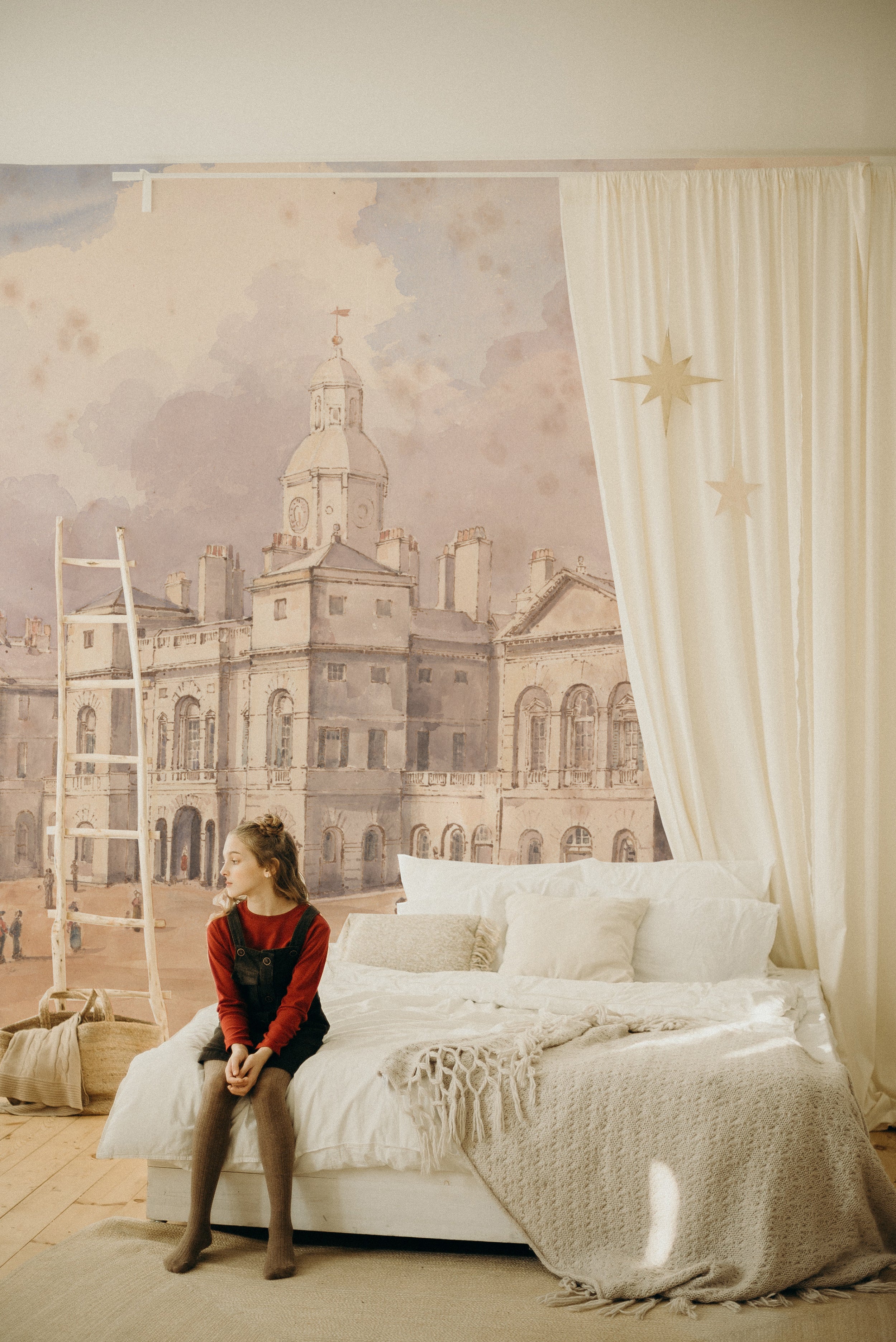 A child's bedroom with a whimsical touch, featuring the "Parade" wallpaper mural as a backdrop. The mural's historic cityscape is paired with simple, cozy bedding and a star-adorned curtain, creating a dreamy atmosphere.